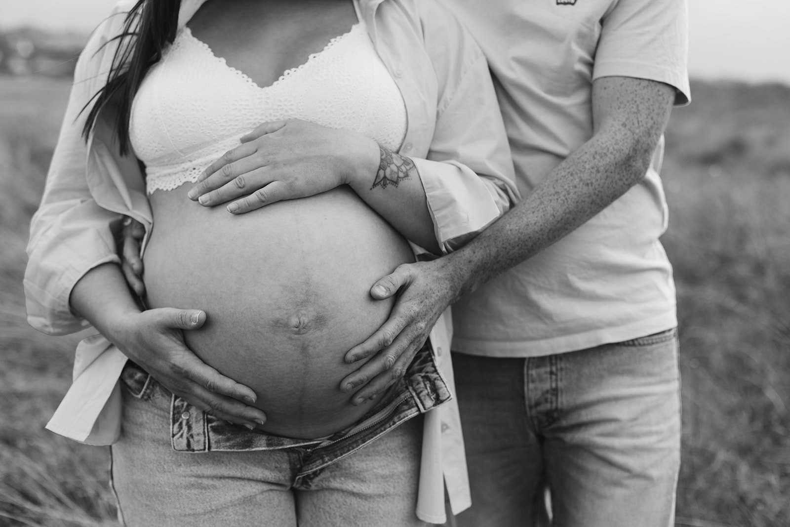Une femme enceinte avec un partenaire posent dans un champ, la main du partenaire repose sur son ventre, en noir et blanc.