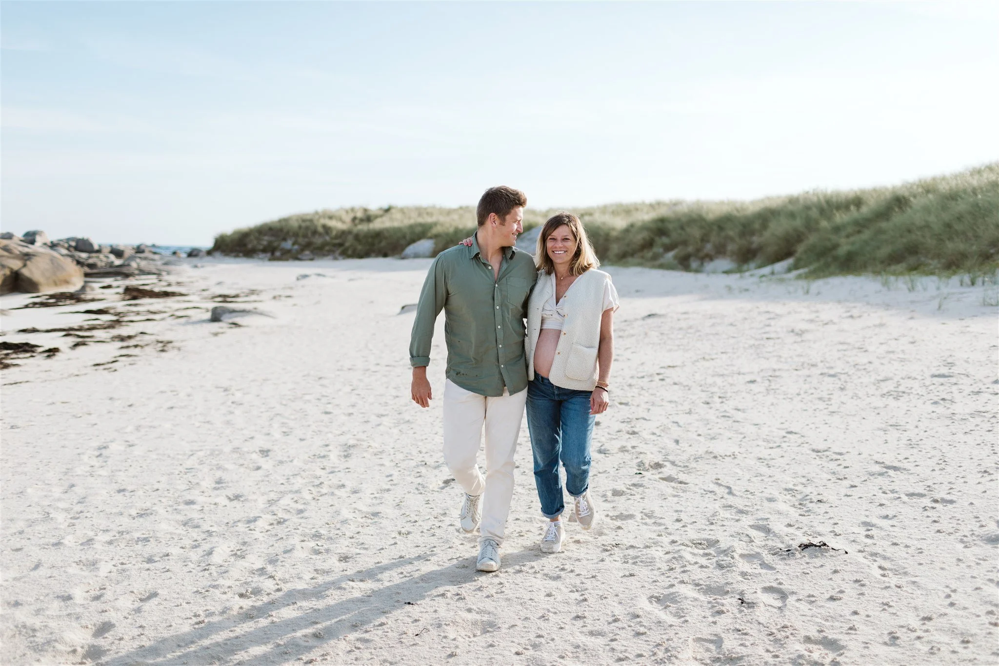 Un couple marche sur une plage avec des dunes de sable et des rochers en arrière-plan, profitant d'une journée ensoleillée.