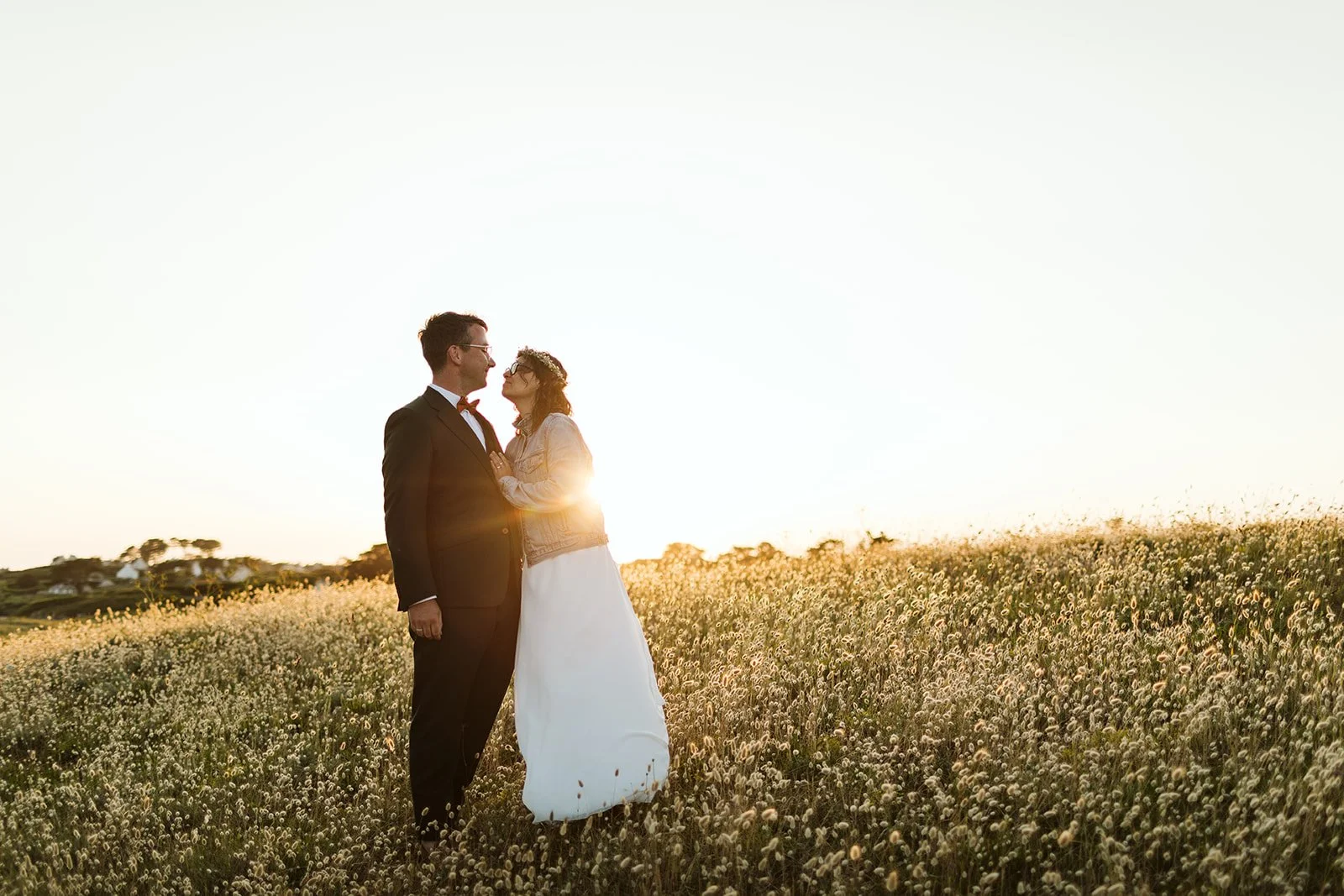 Un couple de mariés se tenant la main dans un champ de fleurs sous un ciel clair, au coucher du soleil.