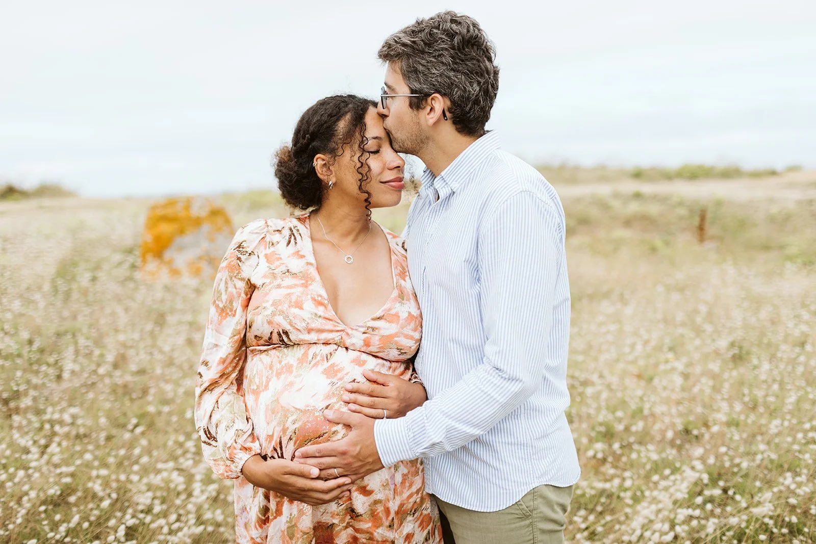 Un couple enceinte se tenant dans un champ de fleurs, l'homme embrassant le front de la femme.
