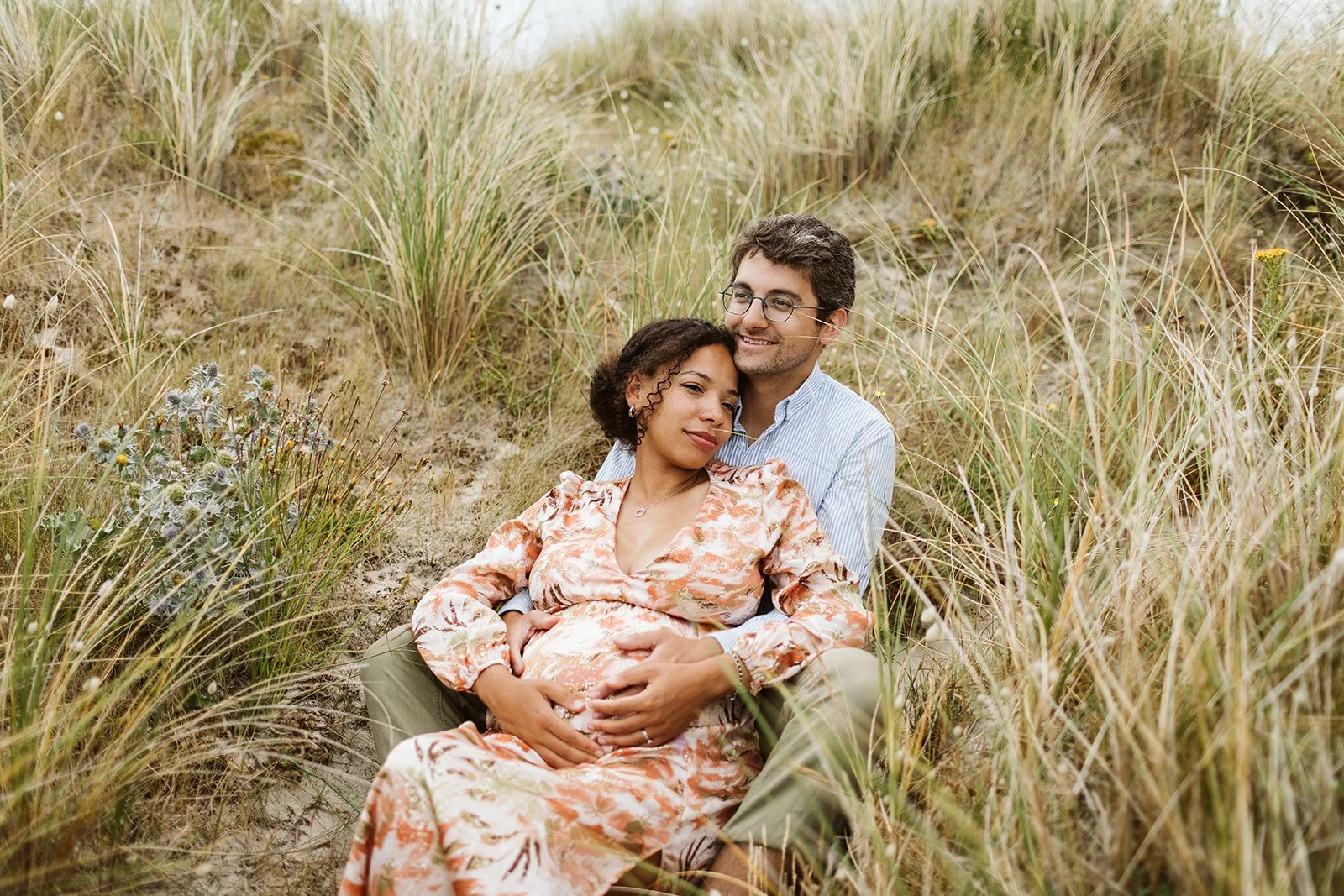 Un couple assis dans la nature, la femme enceinte avec une robe à motifs orange et blanc, l'homme avec une chemise à rayures bleues, entourés d'herbes grasses et de fleurs sauvages.