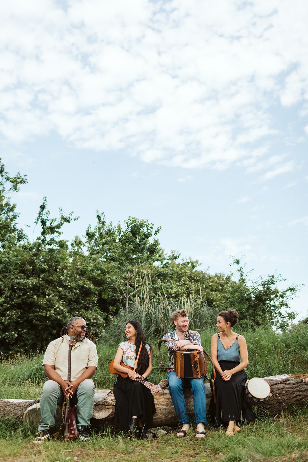 Groupe de quatre personnes assises sur un tronc d'arbre dans un environnement naturel, jouant de la musique et souriant, avec un ciel partiellement nuageux en arrière-plan.