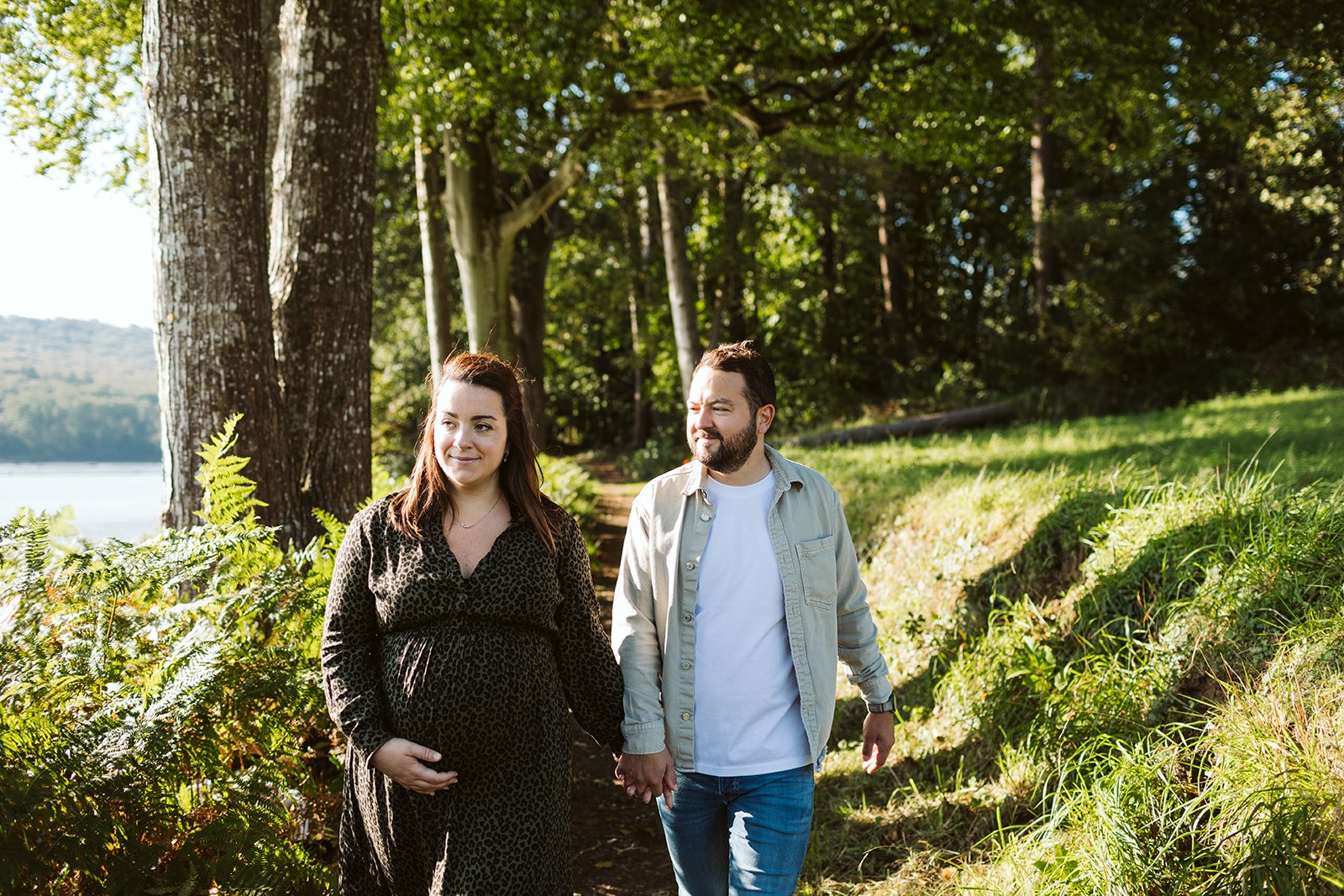 Un couple marche main dans la main dans une forêt ensoleillée, la femme enceinte porte une robe à motifs léopard et l'homme porte une veste légère et un t-shirt blanc.