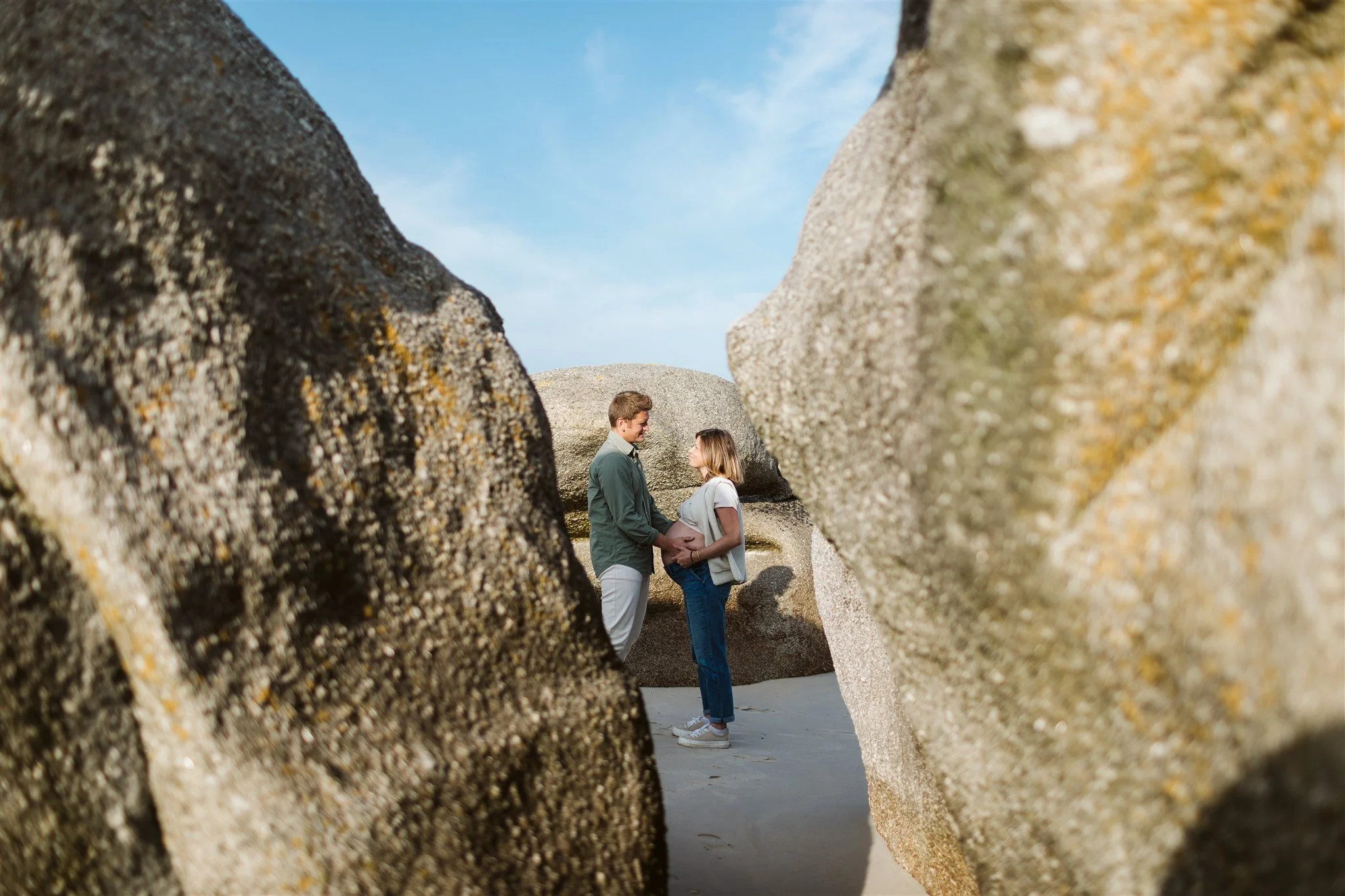 Un couple debout face à face entre deux grandes roches sur une plage, tenant la main et se regardant tendrement sous un ciel bleu.
