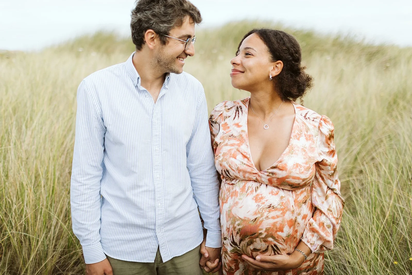 Un couple marche dans un champ de hautes herbes, se tenant la main et se regardant avec douceur, l'homme porte une chemise à rayures et la femme une robe fluide à motifs, elle est enceinte.