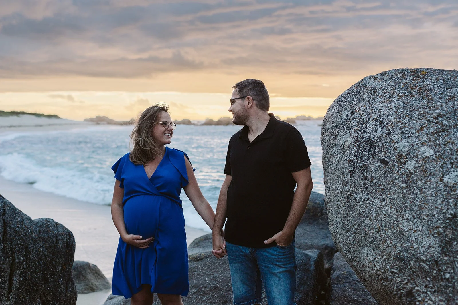 Un couple, une femme enceinte, et un homme, se tiennent par la main sur une plage rocheuse au coucher du soleil.