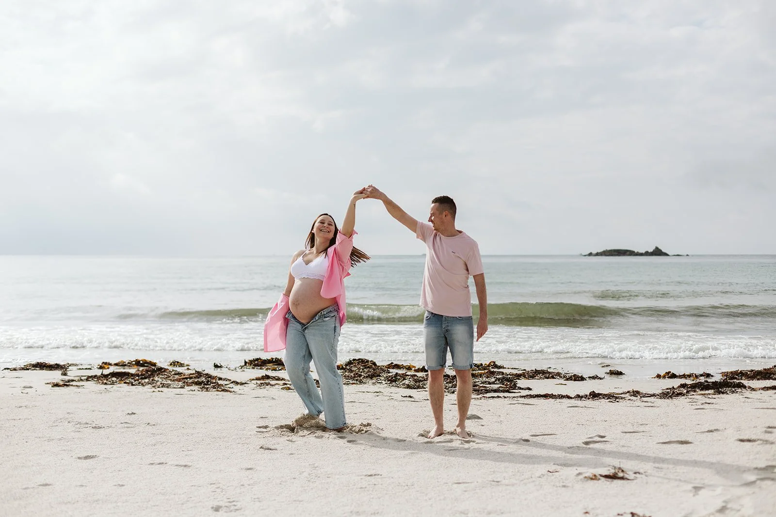 Un couple de jeunes sur la plage, la femme enceinte, danse et joue avec son partenaire qui lui tient la main, en arrière-plan la mer et un ciel nuageux.