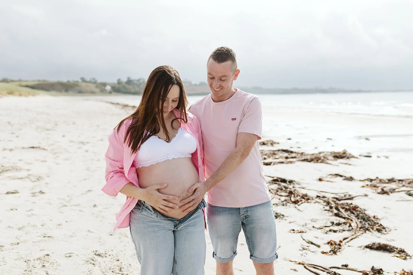 Un couple souriant sur une plage, la femme enceinte portant un haut blanc et une chemise rose, l'homme en t-shirt rose, tenant le ventre de la femme