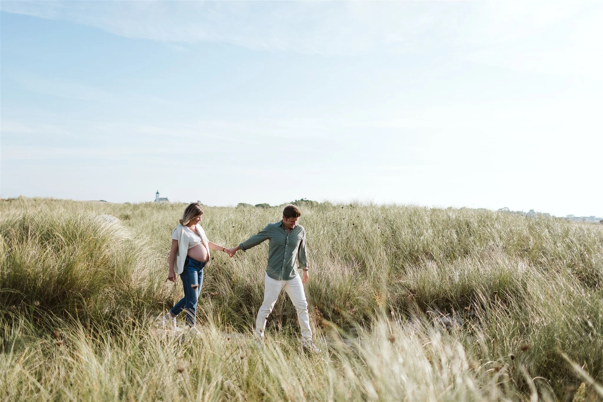 Un couple marche dans un champ d'herbes hautes, tenant la main, dans un paysage ensoleillé avec un ciel bleu clair.