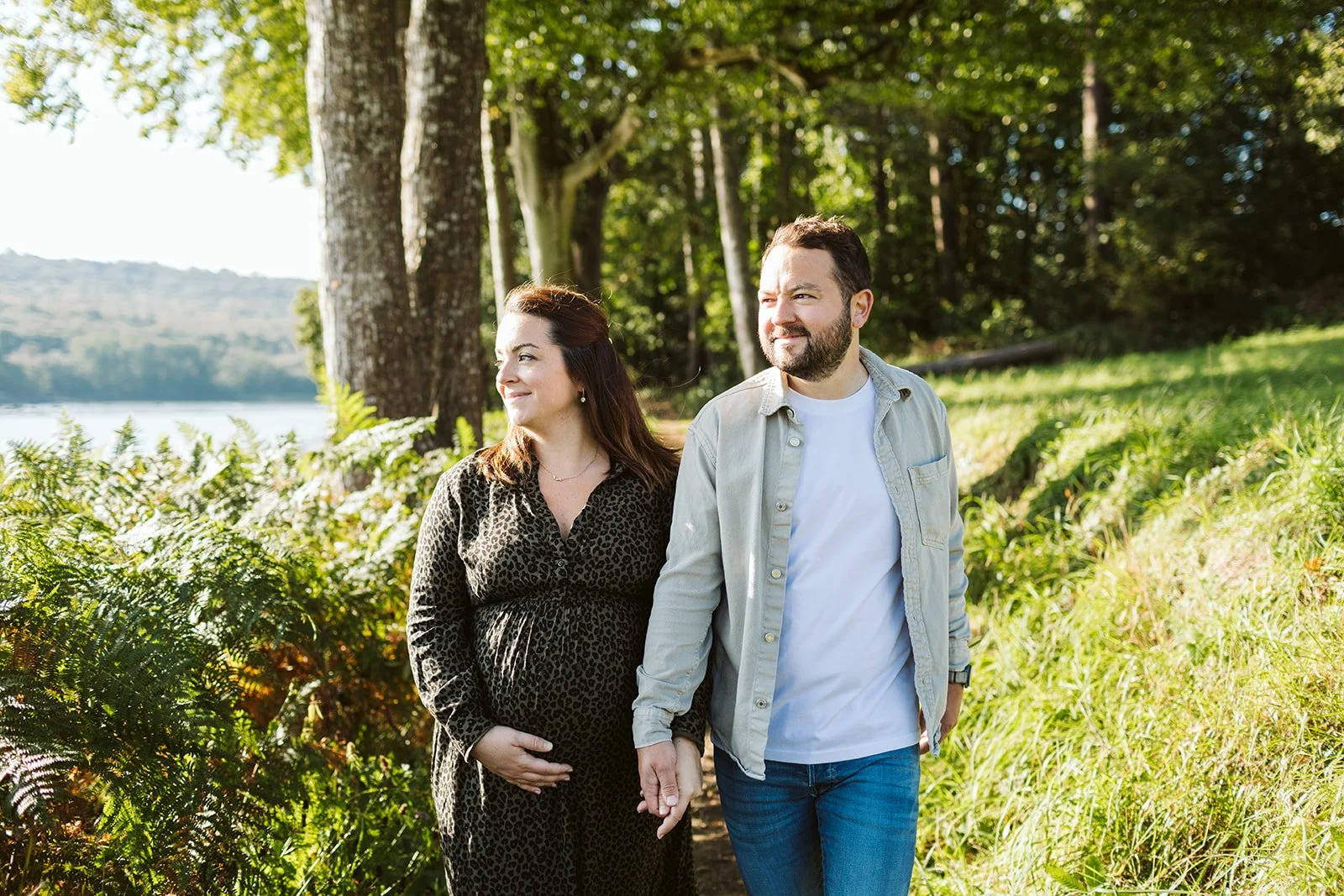 Un couple marchant main dans la main dans un parc entouré d'arbres et de verdure, avec un plan d'eau visible en arrière-plan, sous un ciel ensoleillé.