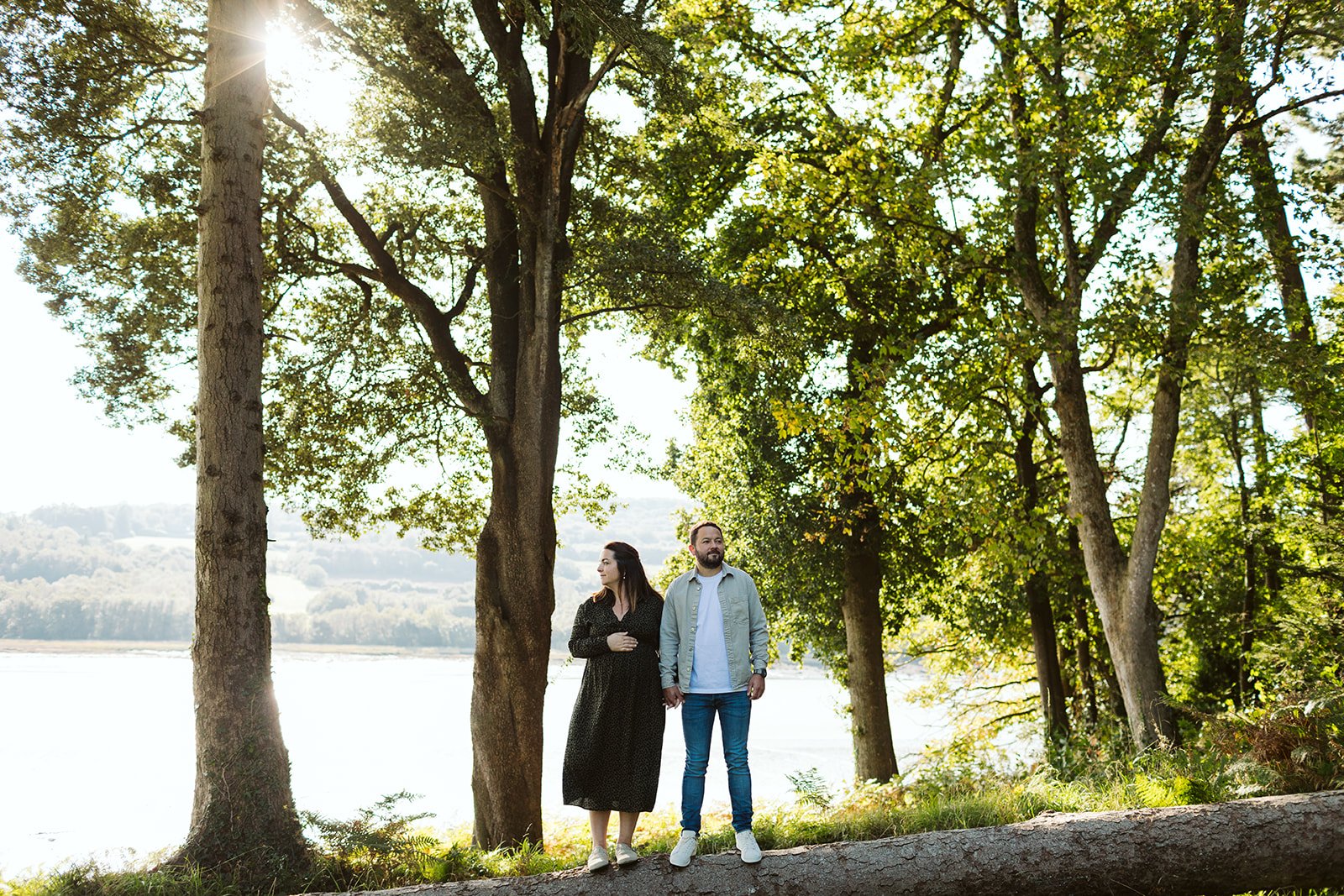 Un couple marche main dans la main dans un parc près de la rivière, entouré d'arbres verts avec le soleil brillant derrière eux.