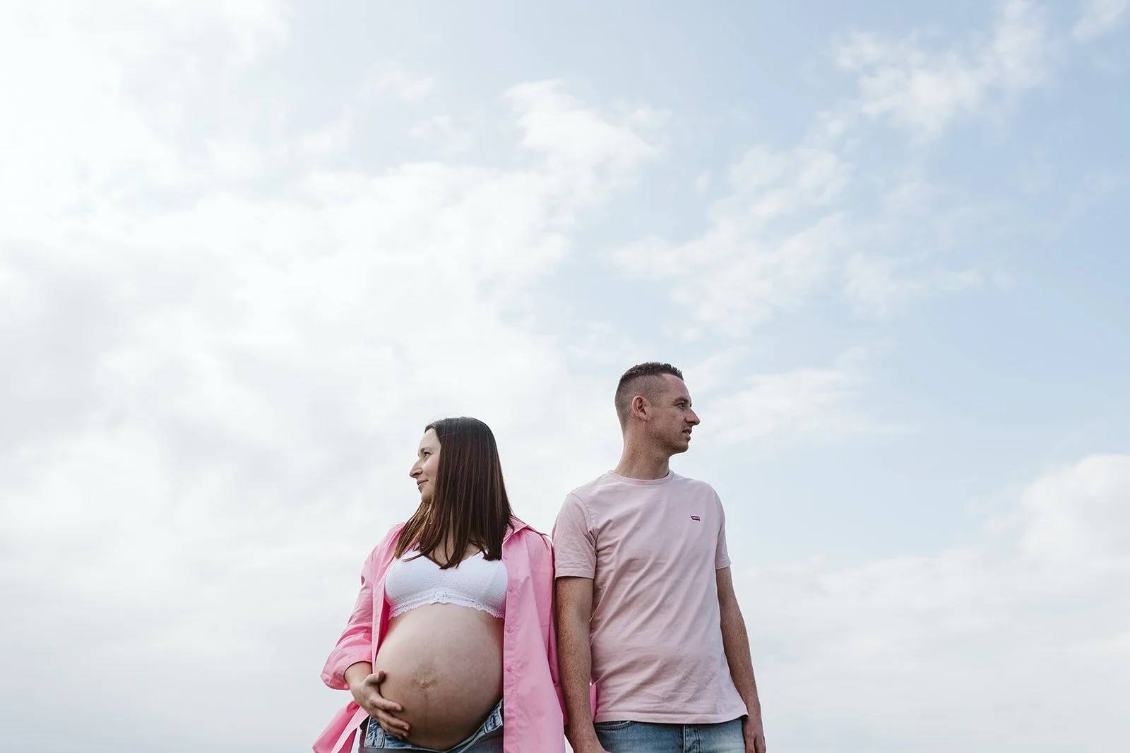 Une femme enceinte et un homme se tiennent dos à dos à l'extérieur par une journée ensoleillée avec un ciel partiellement nuageux.