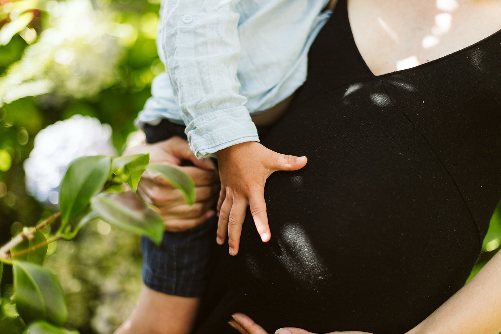 Un enfant tient la main d'une femme, probablement sa mère, dans un cadre extérieur avec de la verdure. La mère porte une tenue noire et il y a de la lumière naturelle.