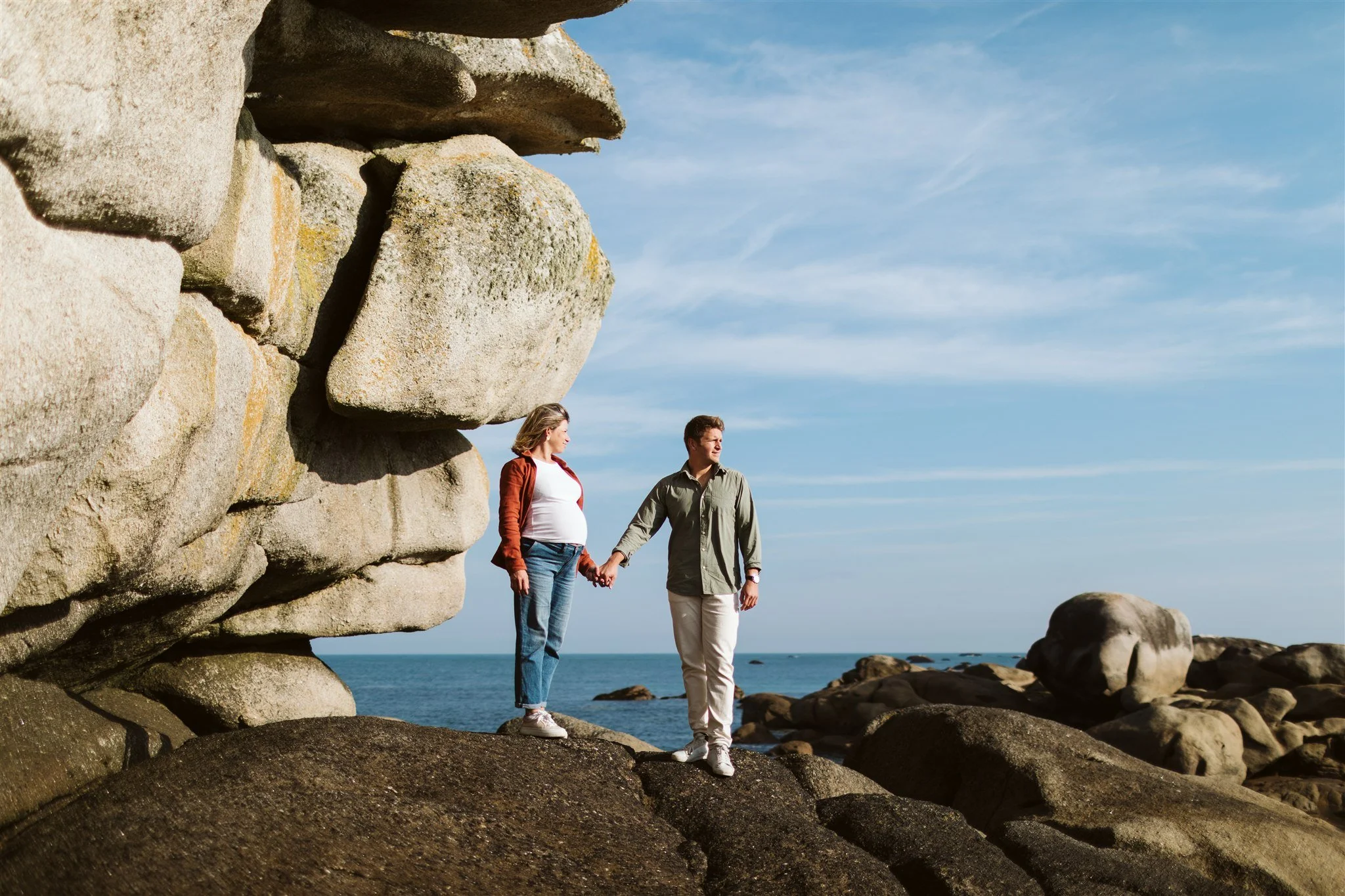 Un couple marche main dans la main sur des rochers au bord de la mer avec un grand rocher en pierre à gauche et un ciel bleu en arrière-plan.