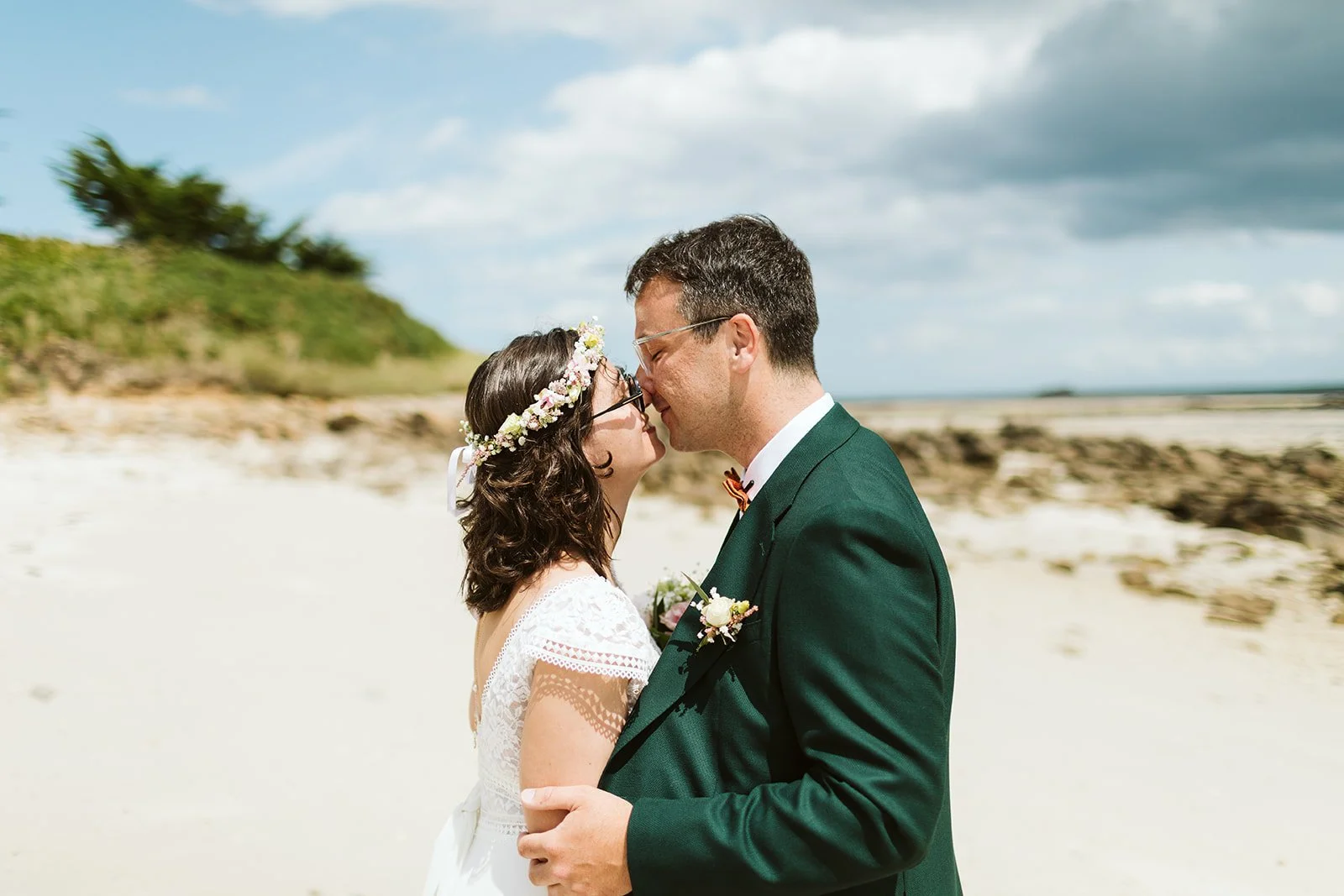 Un couple de mariés s'embrassant sur la plage, la femme porte une couronne de fleurs et une robe blanche, l'homme porte un costume vert avec une broche fleurie.