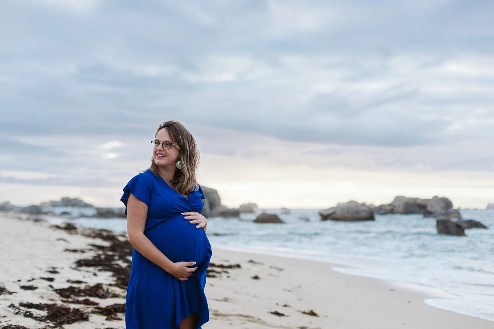 Femme enceinte en robe bleue sur la plage, regardant à gauche, avec des rochers en mer et un ciel nuageux en arrière-plan.