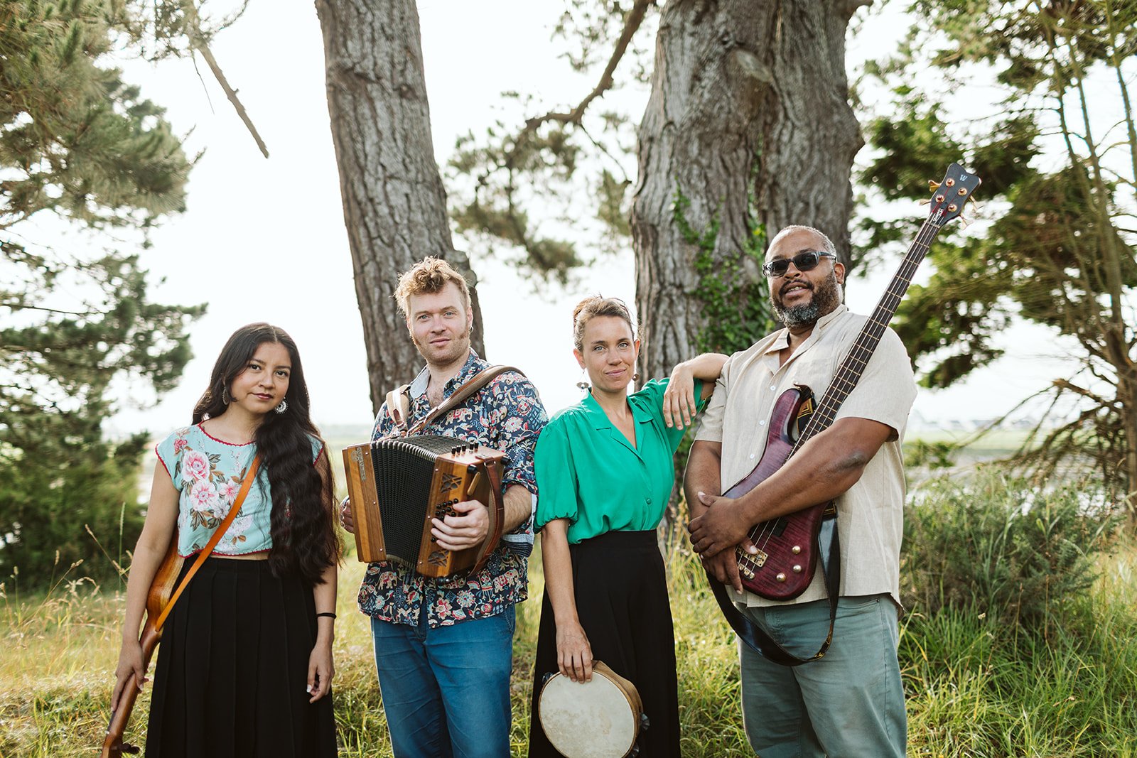 Un groupe de quatre musiciens posant à l'extérieur, entouré d'arbres et d'herbe verte. Ils tiennent différents instruments de musique, dont une guitare, un accordéon, un tambourin, et une guitare basse.