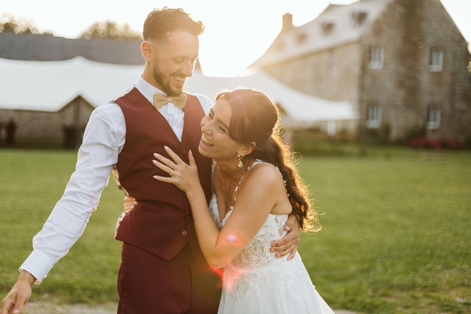 Un couple de mariés souriants, lors de leur mariage en plein air, avec un bâtiment en pierre en arrière-plan et le soleil de fin de journée qui brille.