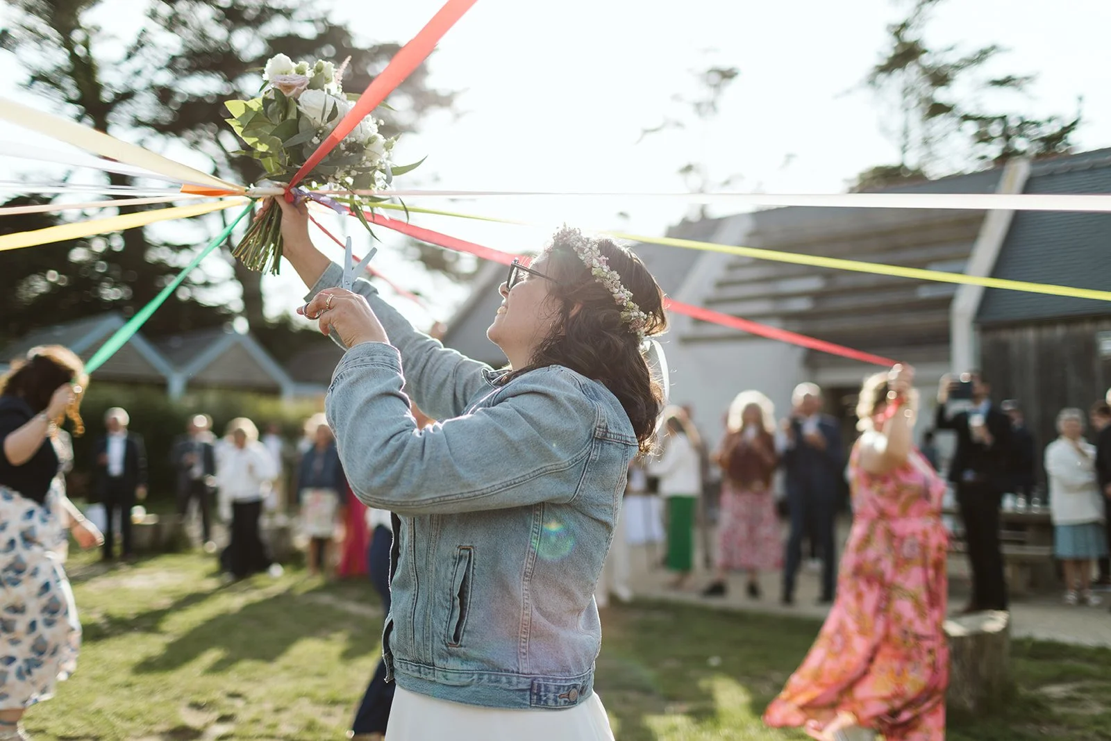 Une femme en jean avec une couronne de fleurs coupe un bouquet de fleurs lors d'une fête en plein air, avec d'autres personnes en arrière-plan.