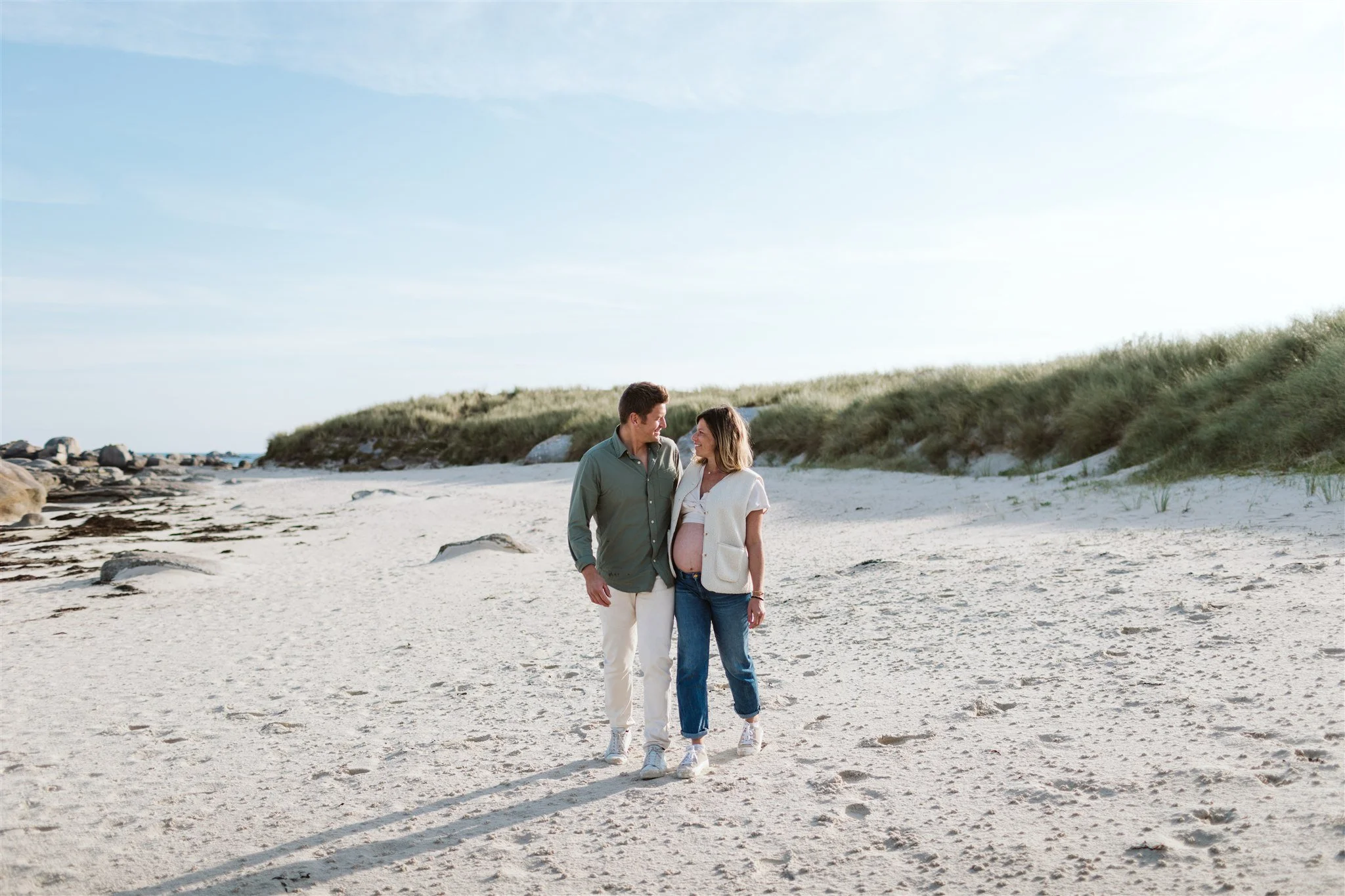 Un couple voyageant le long d'une plage avec une femme enceinte, avec des dunes de sable et un ciel clair en arrière-plan.