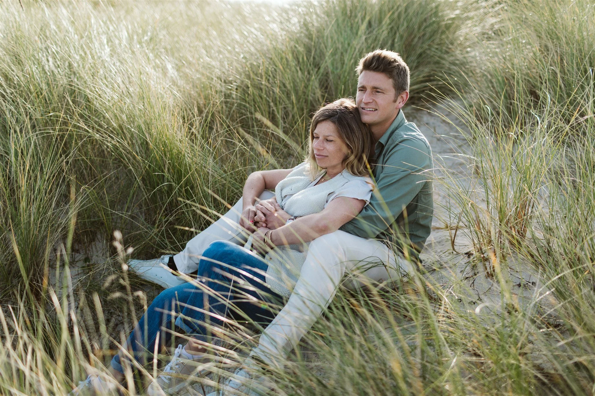Un couple assis dans une dune de sable entourée d'herbes hautes, profitant d'un moment de tendresse.