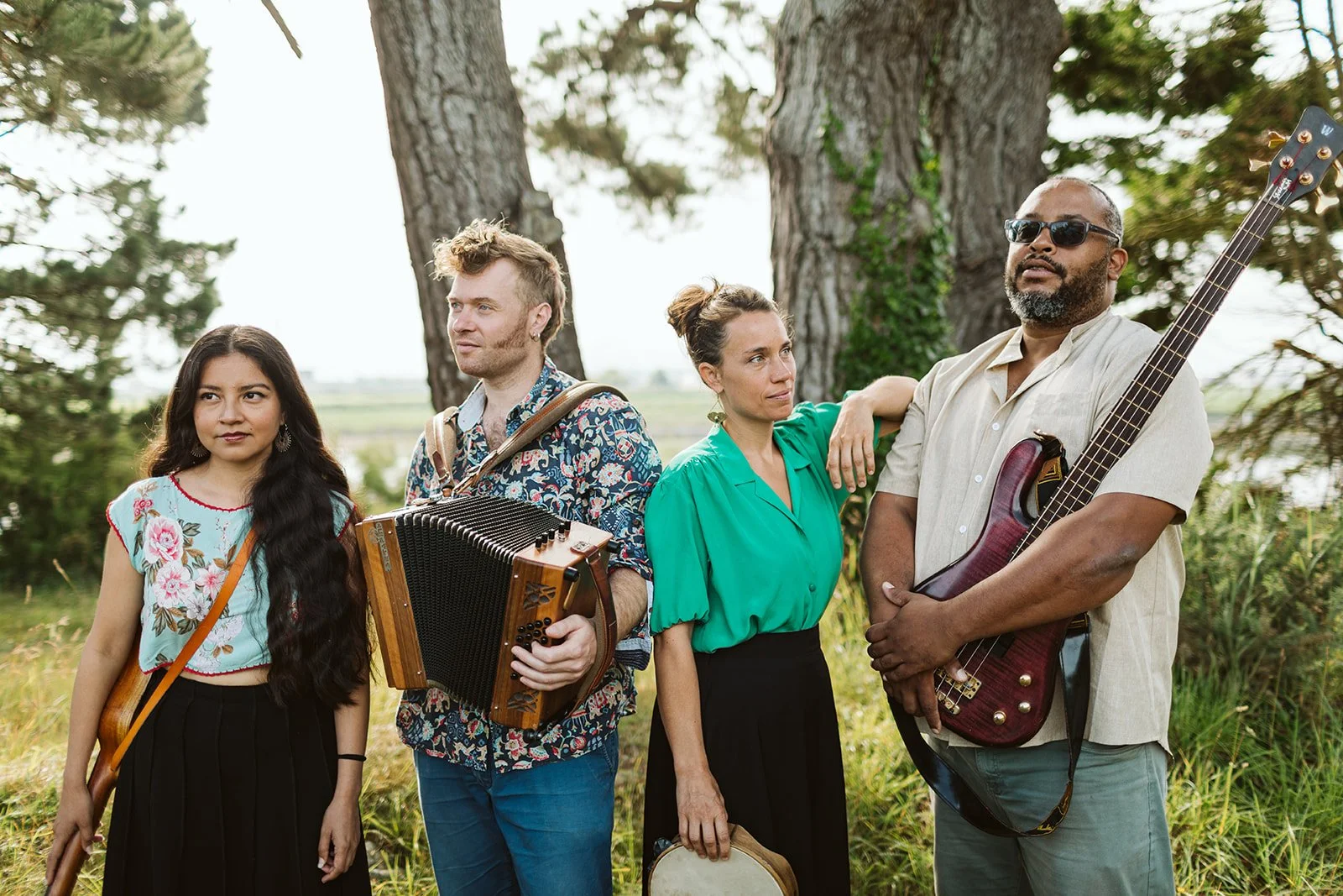 Un groupe de quatre musiciens se tenant à l'extérieur près de grands arbres, avec un champ en arrière-plan, certains portant des instruments de musique.