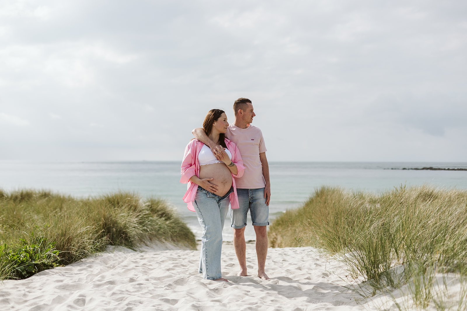 Un couple se promène sur une plage avec une femme enceinte, entouré de dunes et d'une mer calme en arrière-plan.