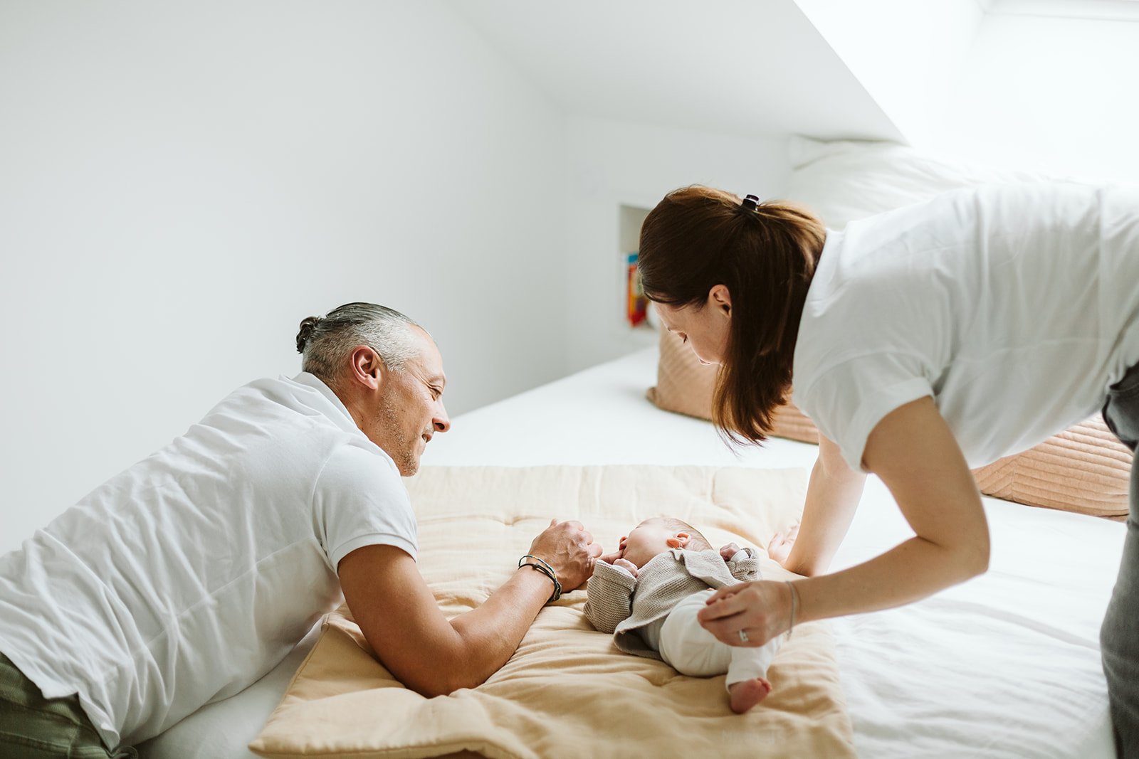 Une famille souriante avec un bébé dans une chambre lumineuse, la mère et le père jouent avec le bébé sur le lit.