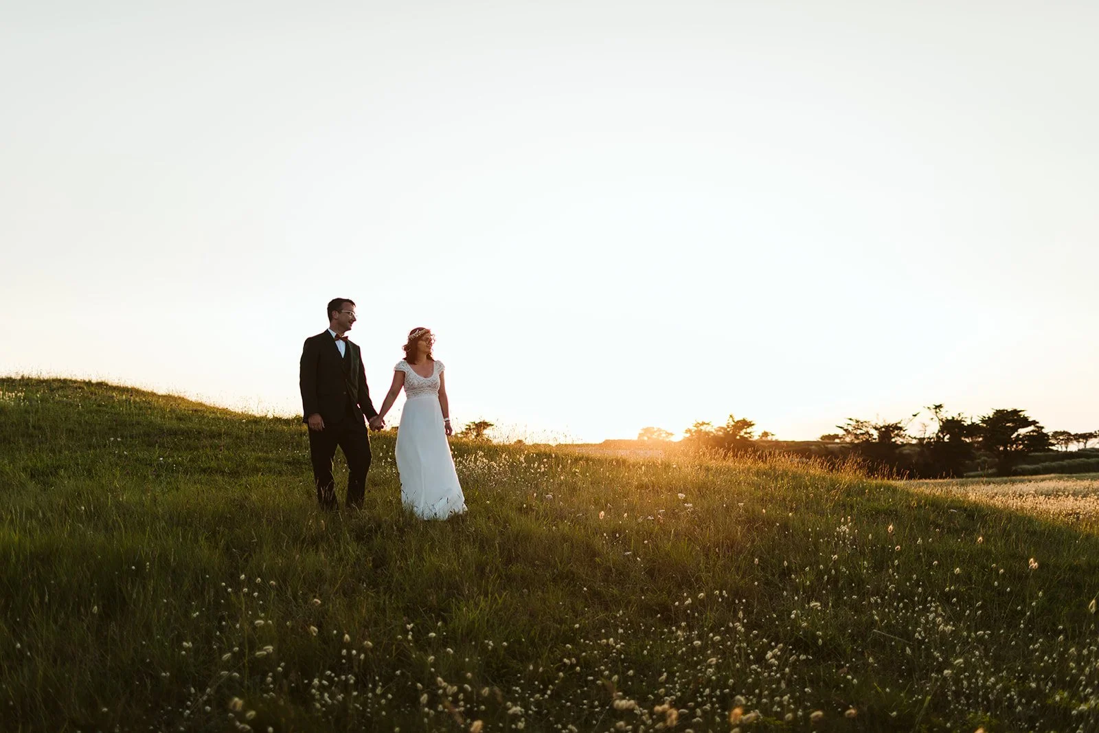 Un couple marié marche dans un champ situé au coucher du soleil, en portant des vêtements de mariage. Le paysage est verdoyant avec des arbres à l'horizon.