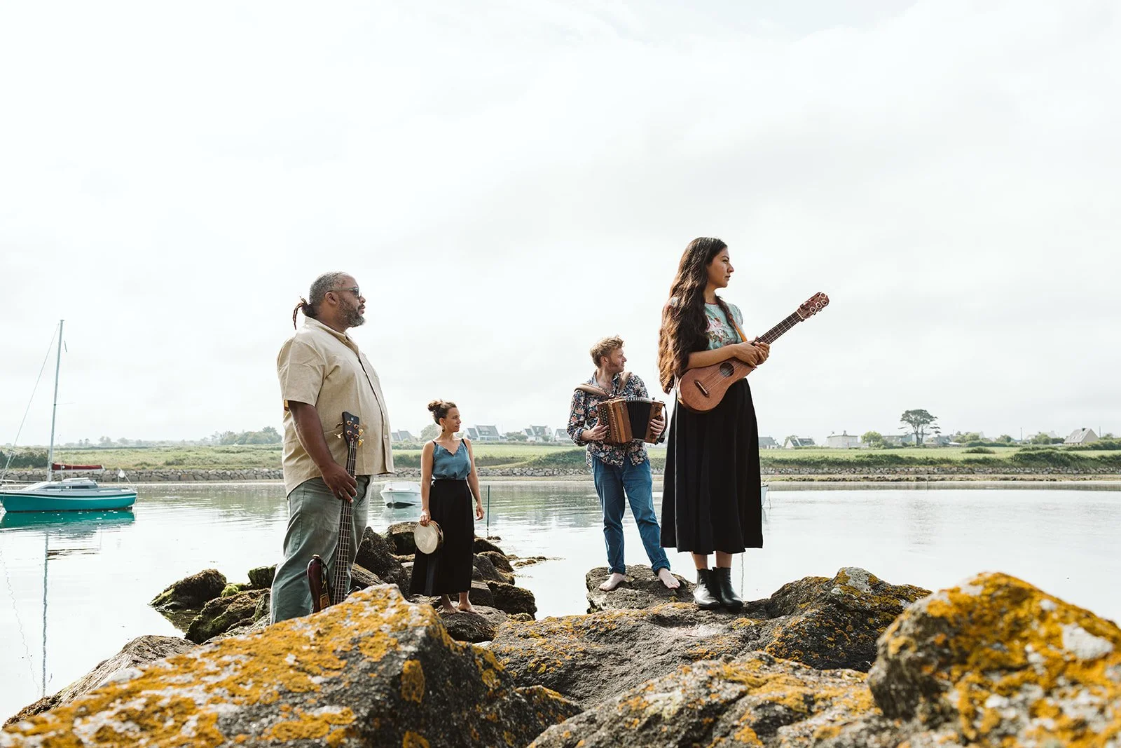 Groupe de cinq musiciens jouant de la musique sur des rochers au bord de l'eau, avec des voiliers et des maisons en arrière-plan, par temps nuageux.