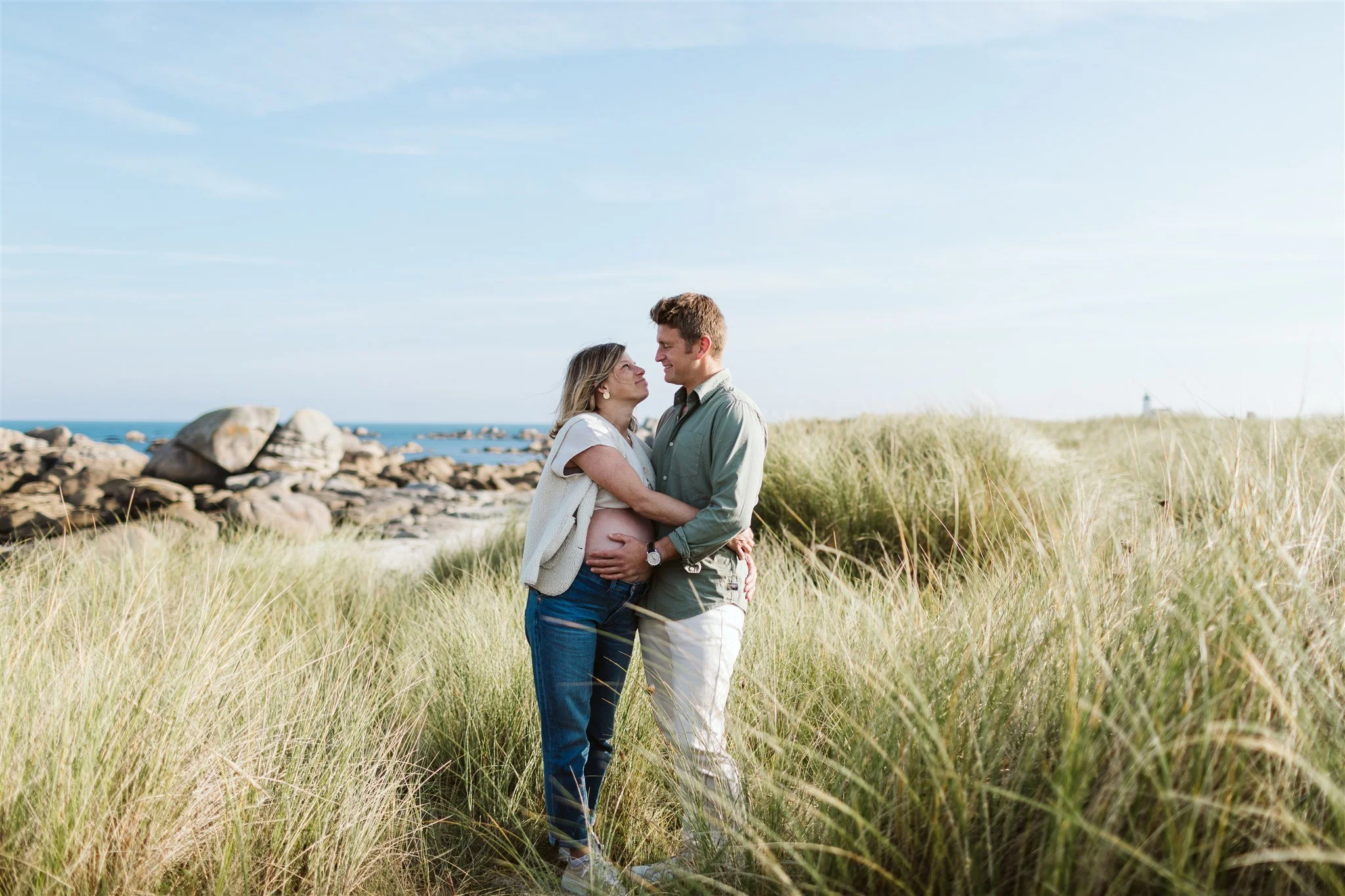 Un couple enceinte se tient dans un champ de hautes herbes près de la mer, échangeant un regard affectueux sous un ciel bleu clair.