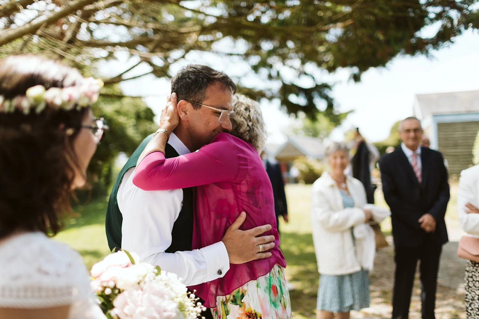 Deux personnes s'embrassent lors d'une cérémonie en plein air, entourées d'autres personnes. La scène se déroule sous un arbre avec des bâtiments en arrière-plan. Il y a des fleurs au premier plan.