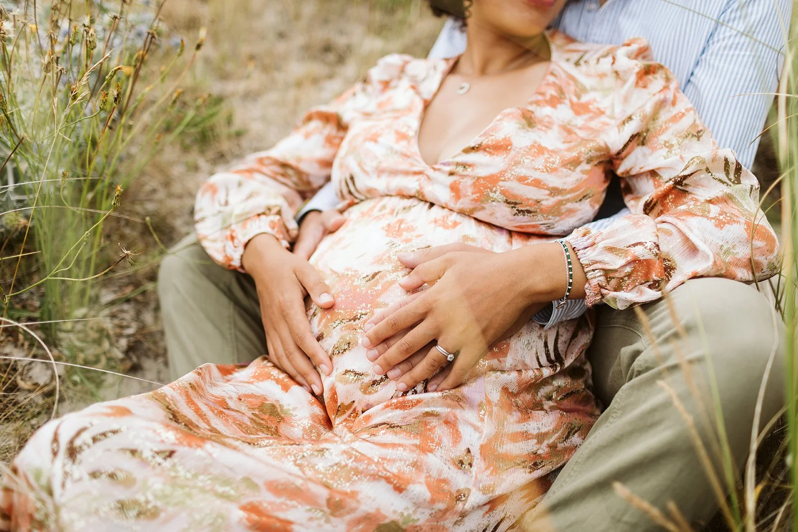 Une femme enceinte allongée sur les genoux de son partenaire dans un champ, main sur son ventre. La femme porte une robe colorée et le partenaire porte une chemise à rayures.