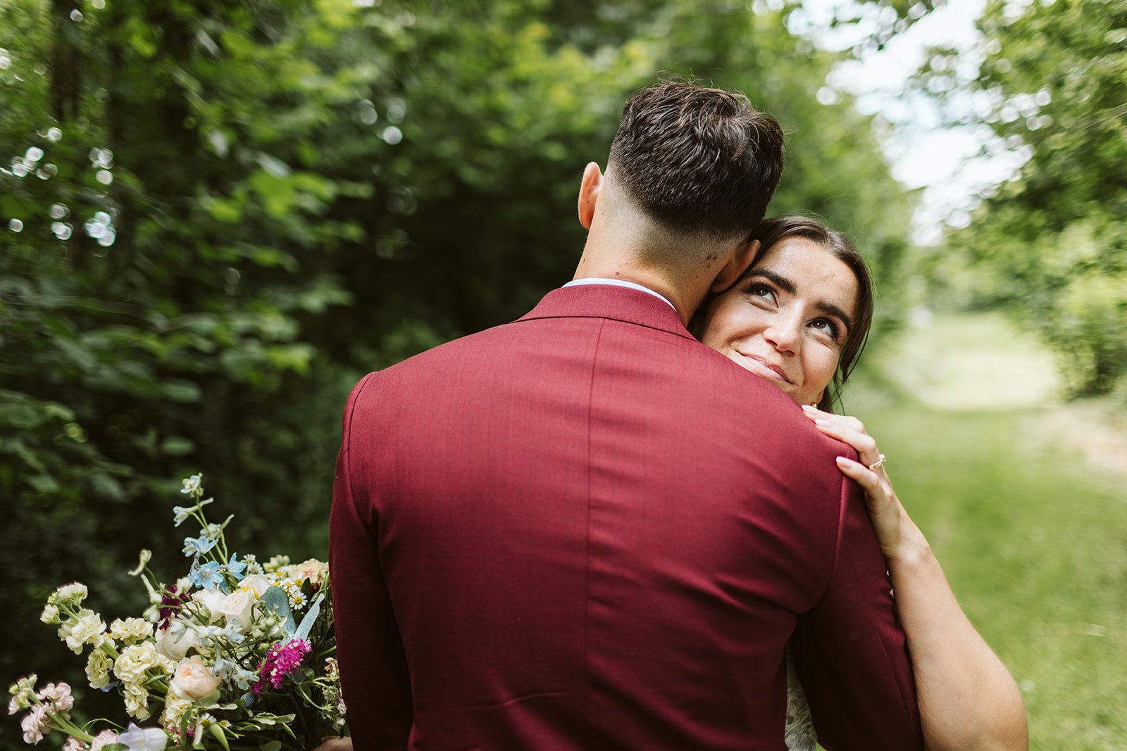 Un couple qui s'étreint dans la nature avec des arbres verts et un ruisseau en arrière-plan, la femme regarde avec un sourire affectueux.