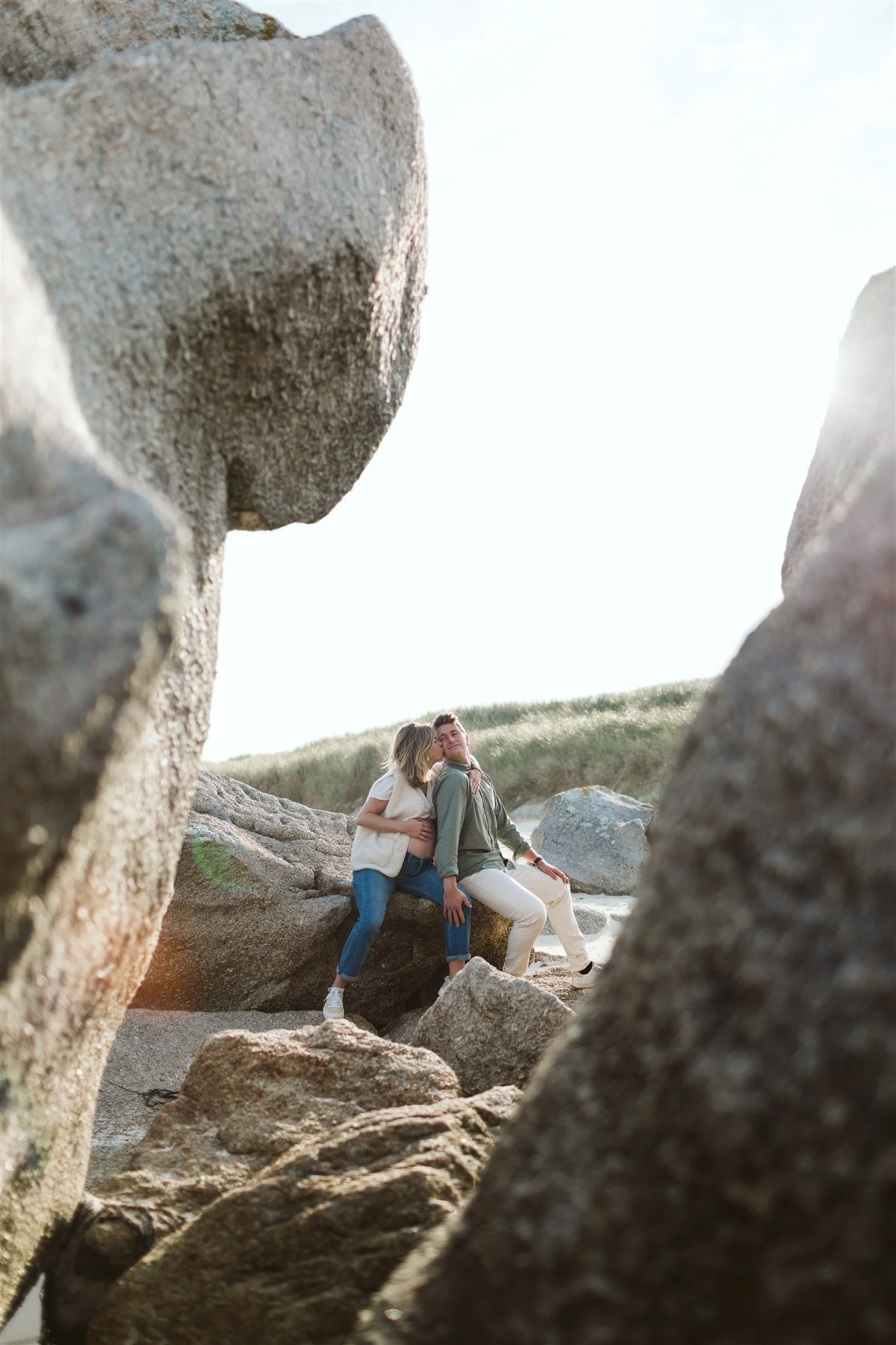 Deux personnes assises sur des rochers à la plage, entourées de grosses pierres, avec un ciel clair en arrière-plan.