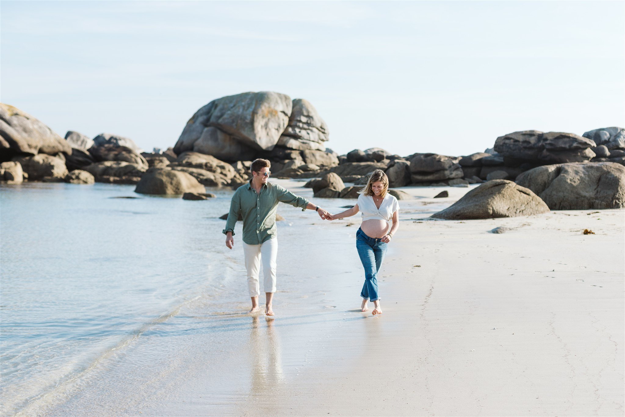 Une femme enceinte et un homme marchent main dans la main sur une plage de sable, avec des rochers en arrière-plan, par une journée ensoleillée.