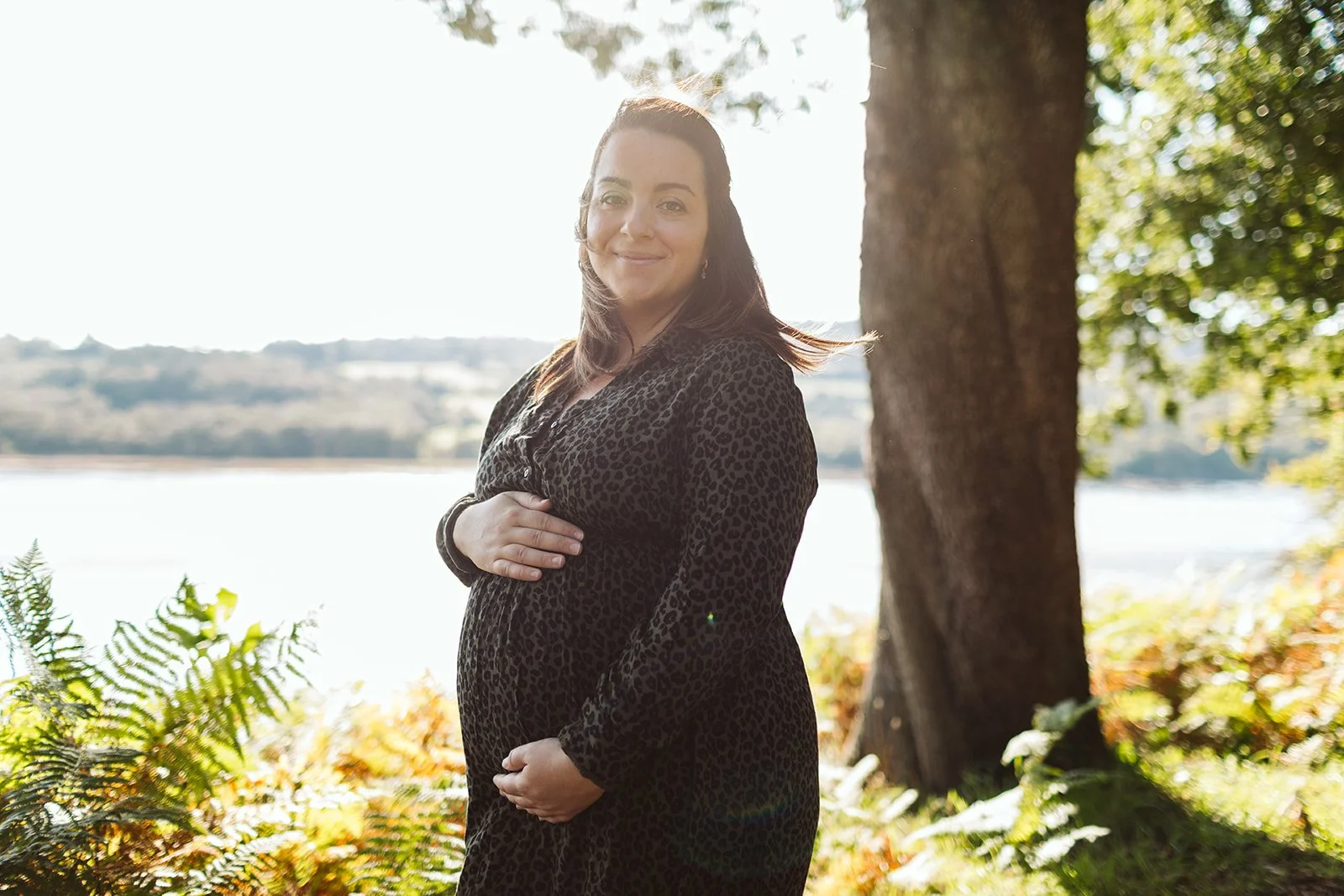 Femme enceinte portant une robe à motif léopard, se tenant le ventre avec une main, en plein air près d'un arbre, avec un lac en arrière-plan et des feuillages verts autour.