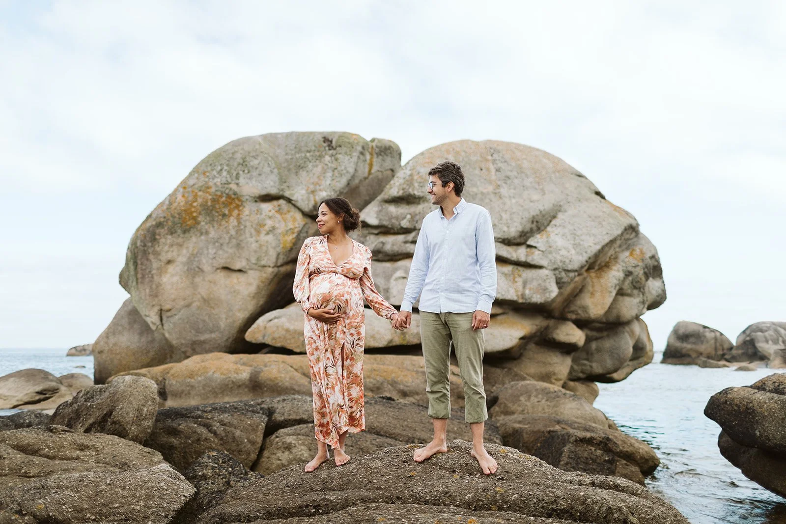 Une femme enceinte et un homme tenant la main se tenant sur des rochers au bord de la mer, avec de gros rochers en arrière-plan.