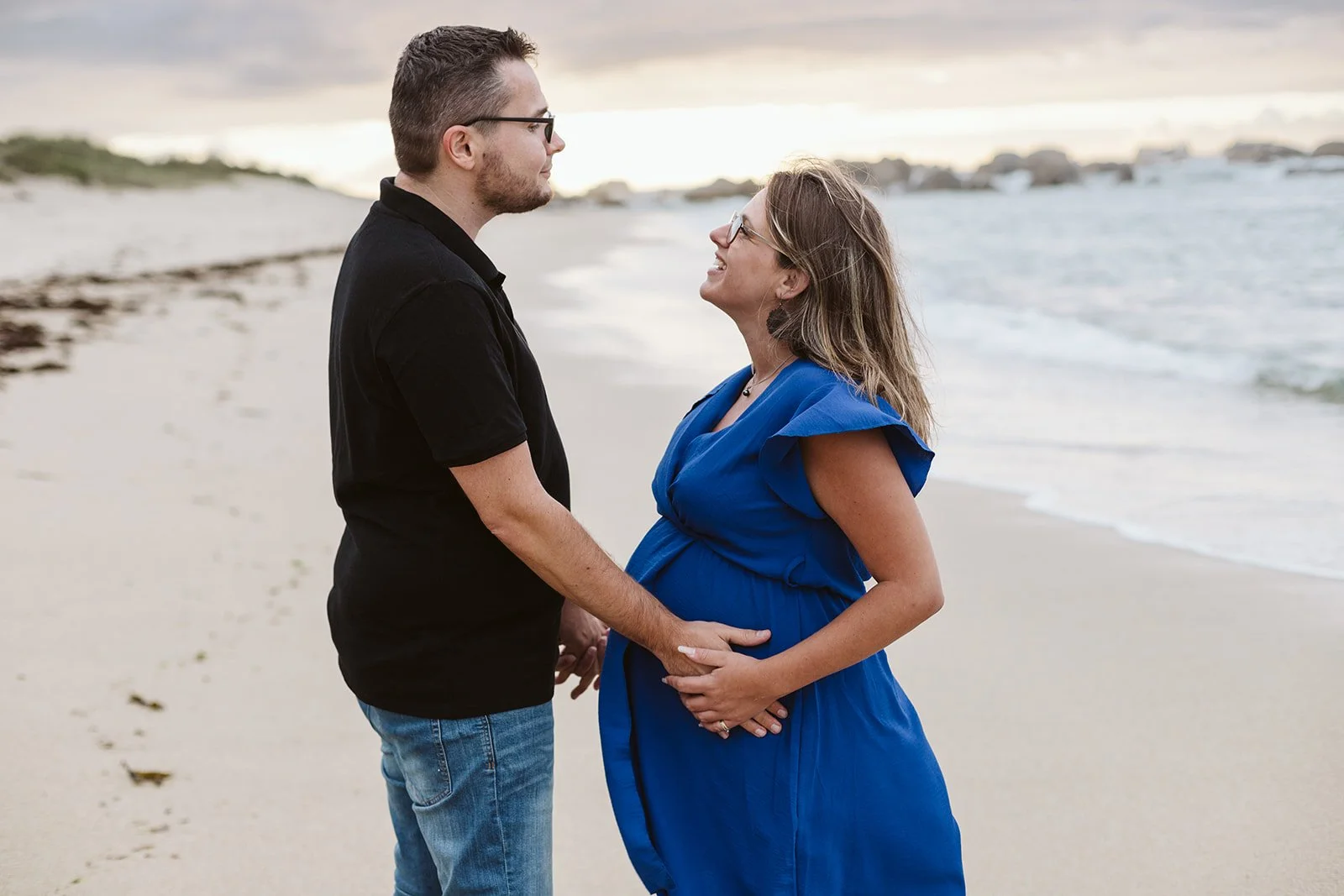 Un couple à la plage, l'homme tenant le ventre de la femme enceinte, ils se regardent avec tendresse.