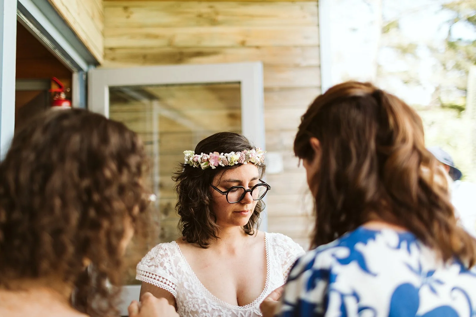 Une femme portant une couronne de fleurs et des lunettes, entourée de deux autres personnes, dans un cadre rustique en bois, probablement lors d'un mariage ou d'une cérémonie en plein air.