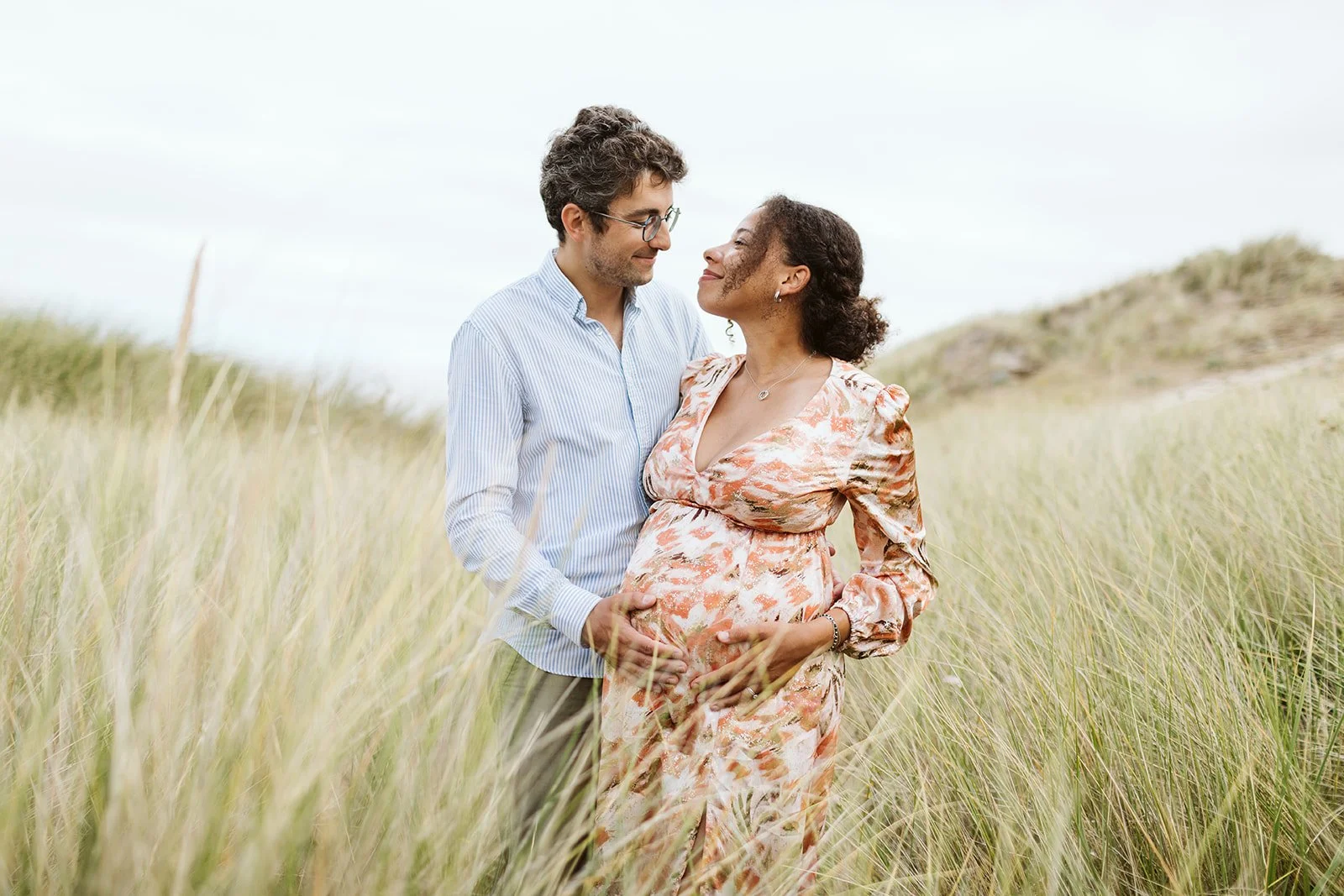 Un couple homme et femme devant un champ de hautes herbes, l'homme tient le ventre de la femme enceinte, ils se regardent tendrement.