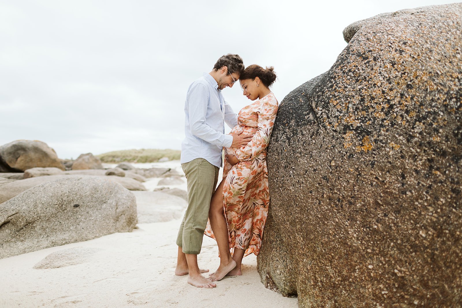 Un couple se tenant la main et se regardant tendrement sur une plage rocheuse, la femme enceinte appuyée contre un rocher.