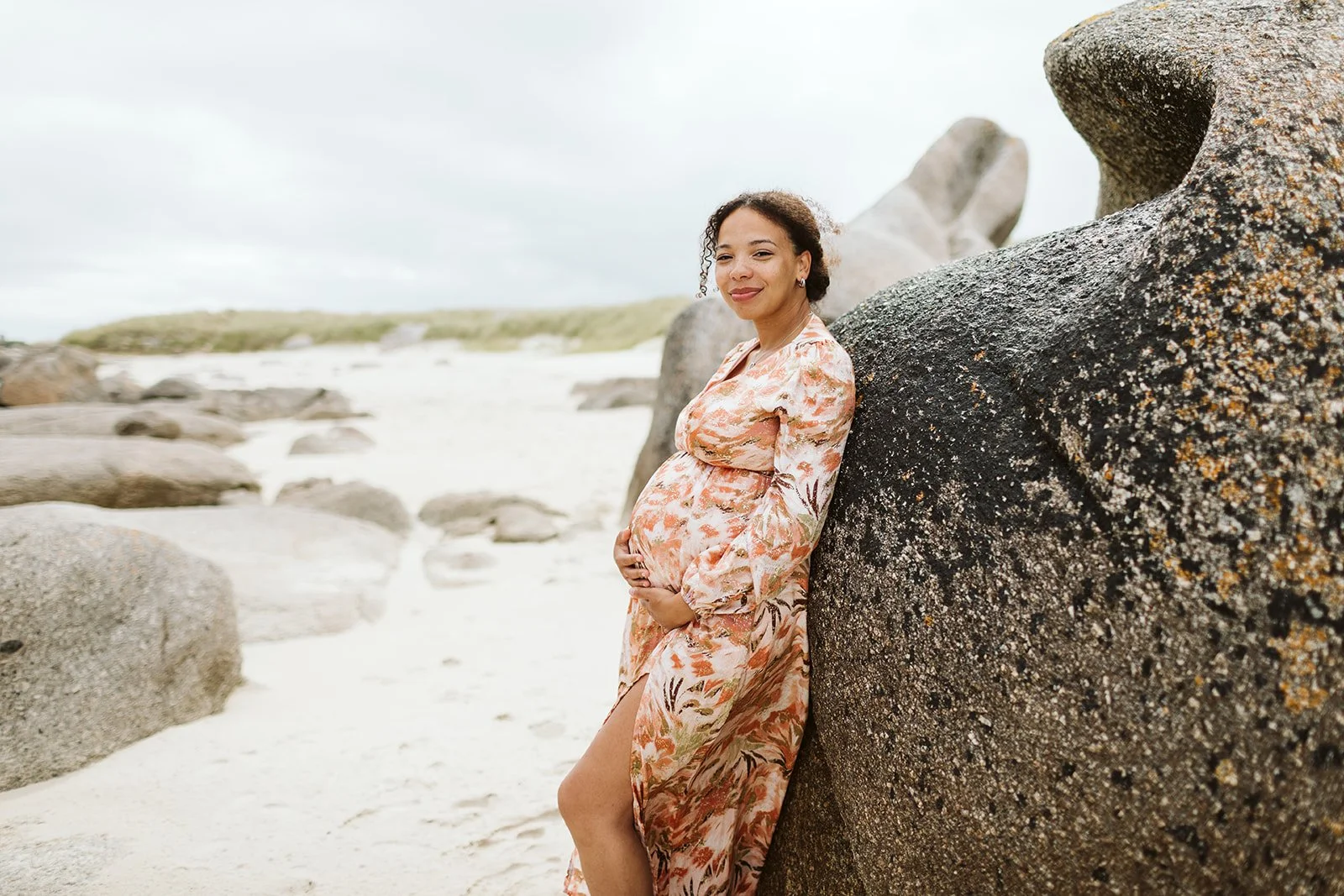 Une femme enceinte souriante portant une robe florale, appuyée contre un gros rocher sur la plage, avec des pierres et du sable en arrière-plan.