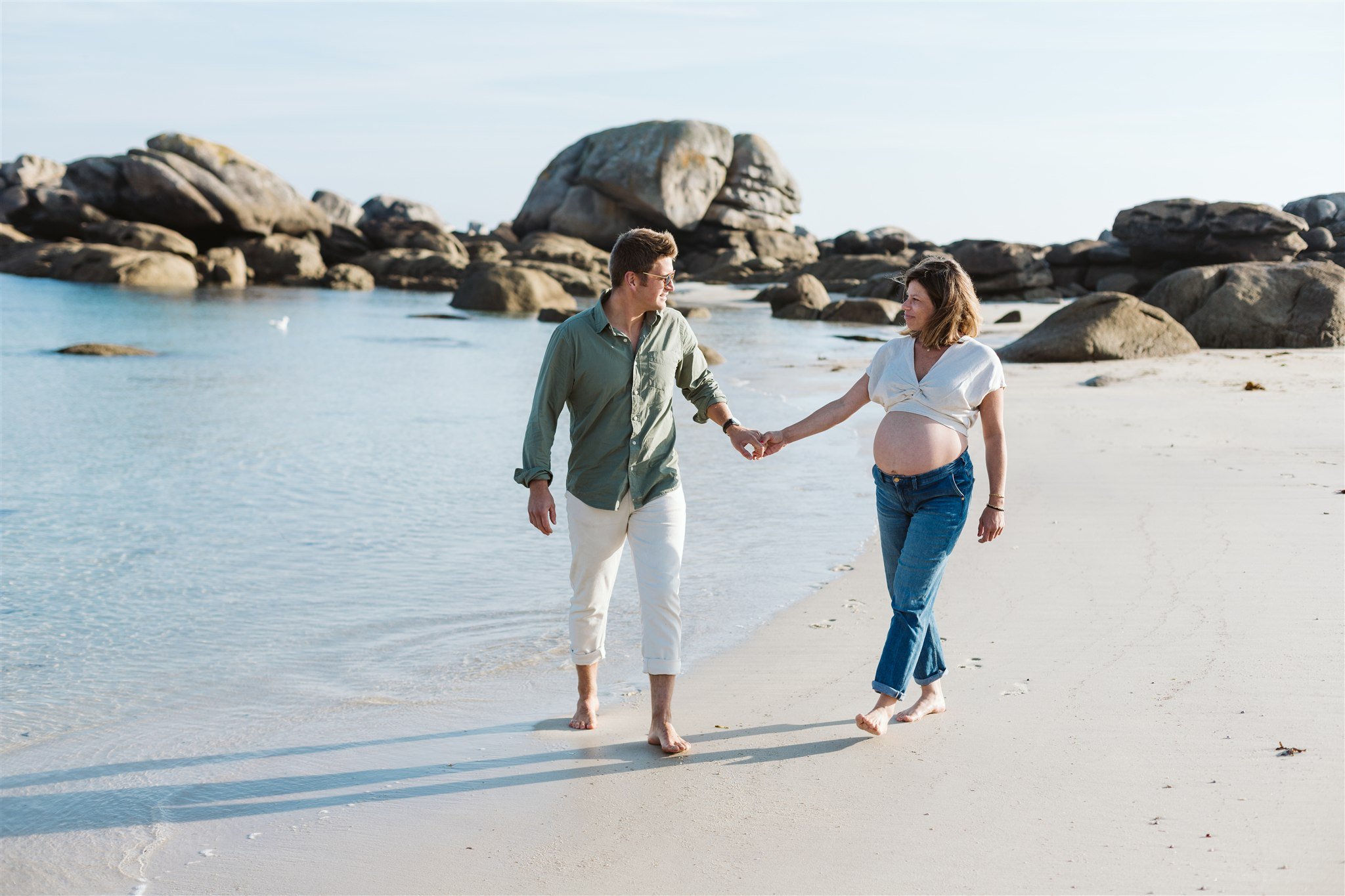 Une femme enceinte et un homme marchent main dans la main sur une plage de sable, avec des rochers en arrière-plan, par une journée ensoleillée.