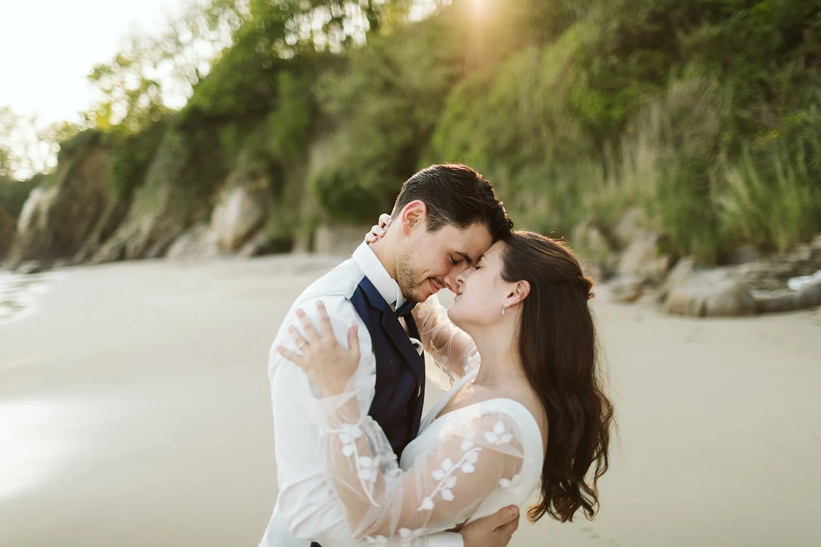 Un couple de mariés embrassés tendrement sur la plage au coucher du soleil avec des falaises verdoyantes en arrière-plan.
