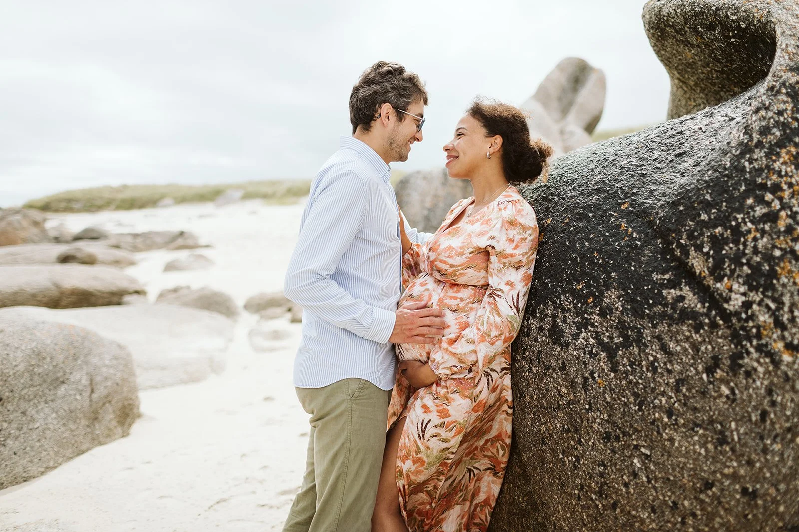 Un couple regardant et touchant le ventre d'une femme enceinte sur une plage rocheuse avec des gros rochers et le ciel nuageux.