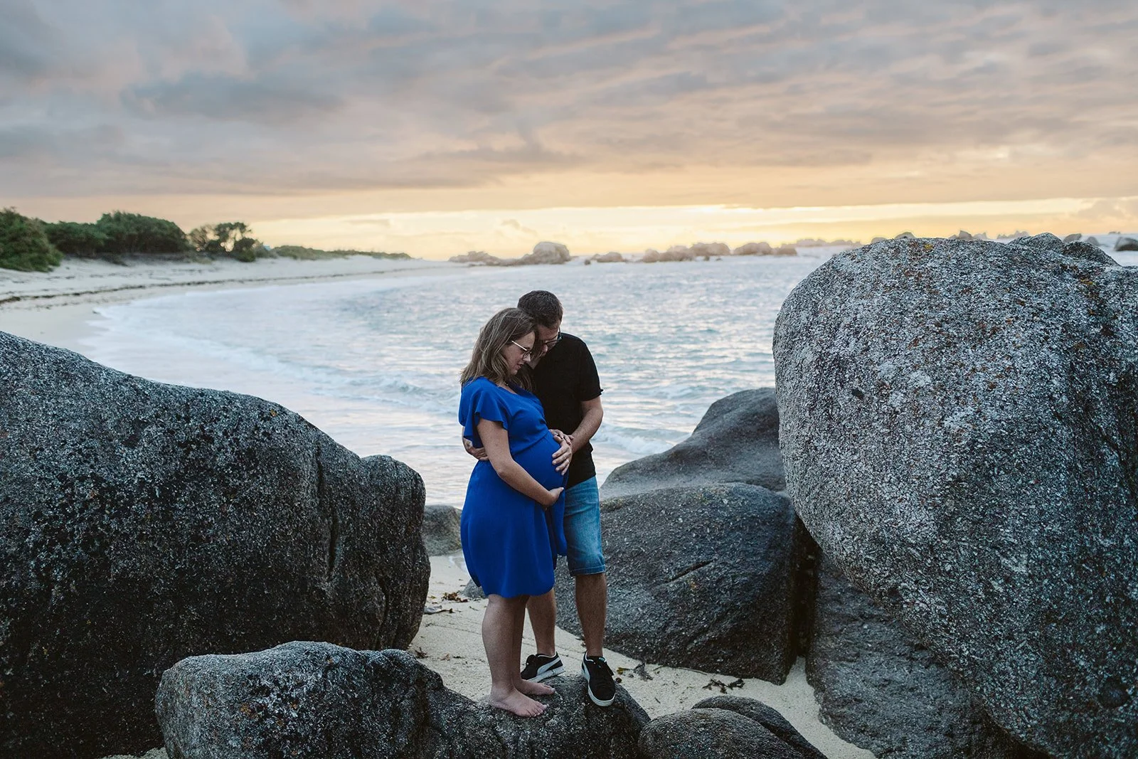Un couple en vacances sur la plage, la femme enceinte, tenant la main de son compagnon, regarde leur ventre. Le coucher de soleil en arrière-plan.