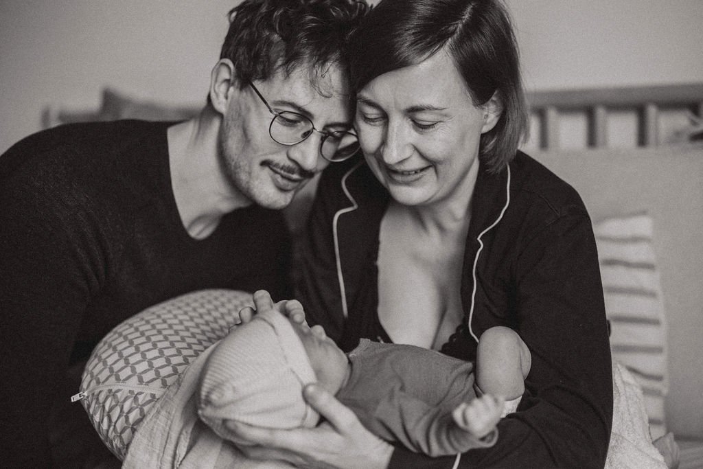 Eltern lächeln ihr Neugeborenes an, Liebe im Wochenbett, dokumentarische Babyfotografie