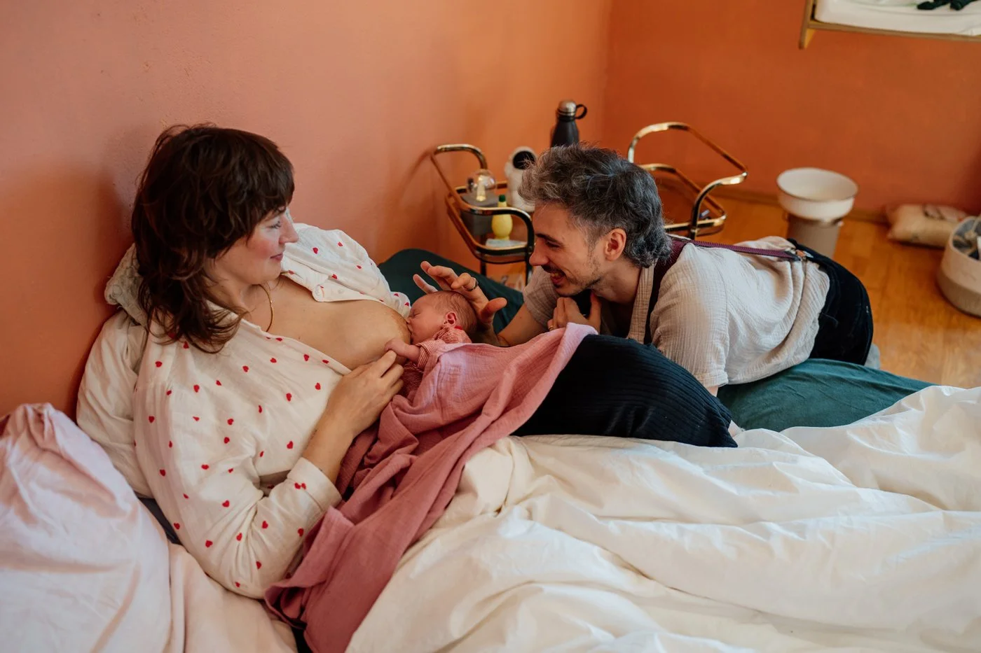 Mama stillt Neugeborenes auf Bett liegend und schaut es liebevoll an während Papa den Kopf streichelt