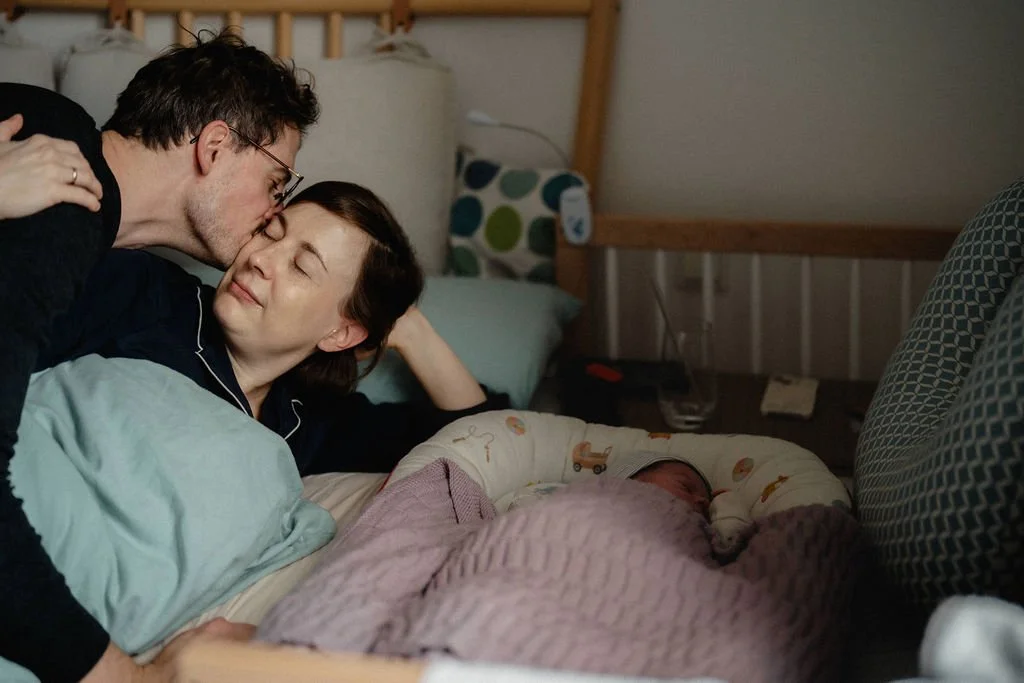 Vater küsst Mutter im Wochenbett, erste Momente als Familie zuhause dokumentiert